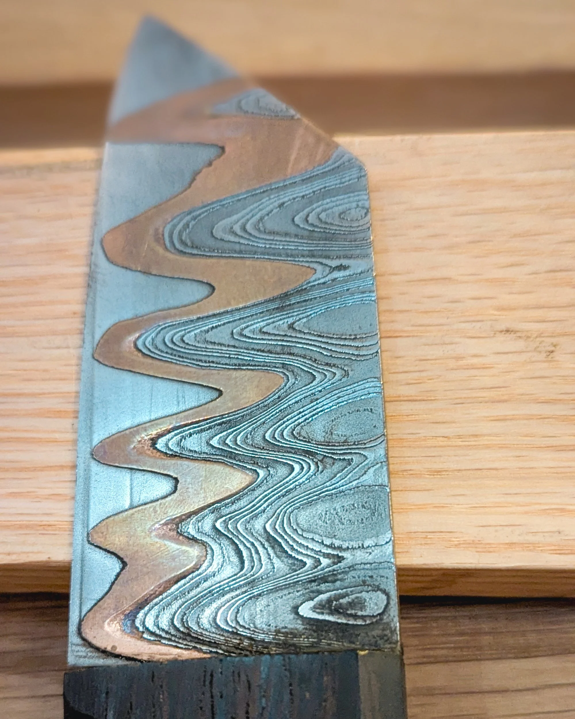 A stunning macro close-up of a Damascus steel knife blade from Oldfield Forge, featuring a prominent, undulating wave of contrasting copper flowing throughout the layered steel pattern.