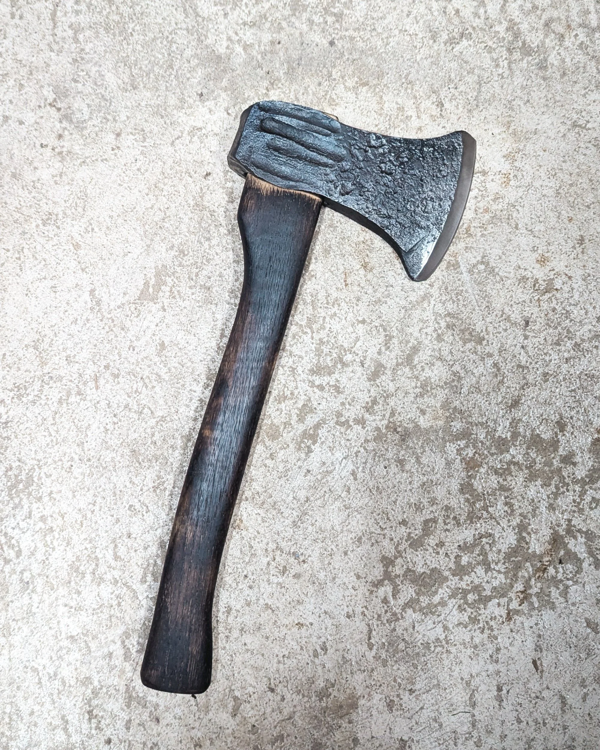 A completed hand-forged axe lying on the rustic floor of a blacksmithing workshop, showcasing the contrast between the dark charred handle and the bright, polished steel blade.