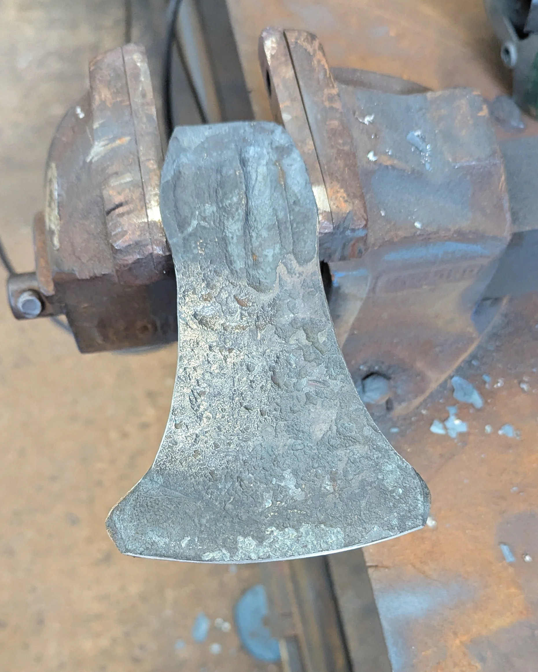 A close-up of a partially finished steel axe head held firmly in a heavy-duty workshop vice, showcasing the rough-forged shape before the final filing and polishing stages.
