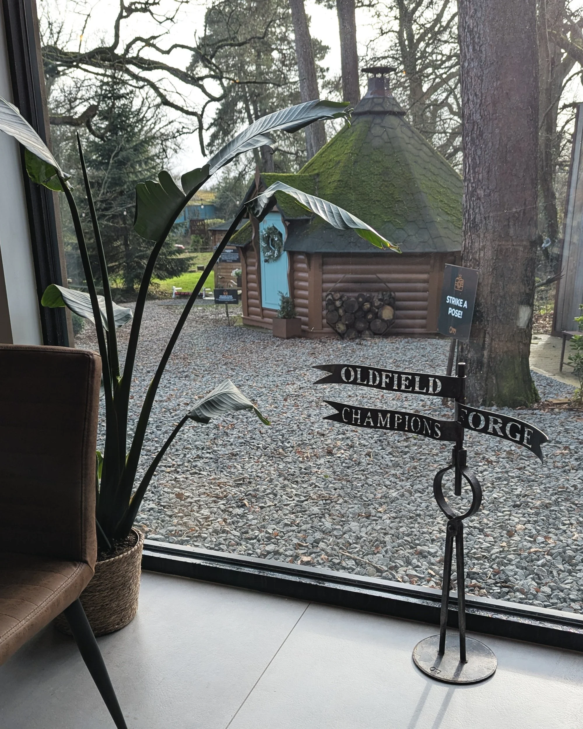 A view looking out from the cafe window at Oldfield Forge, featuring a bespoke freestanding metal brand sign in the foreground and a charming, rustic wooden lodge in the background.