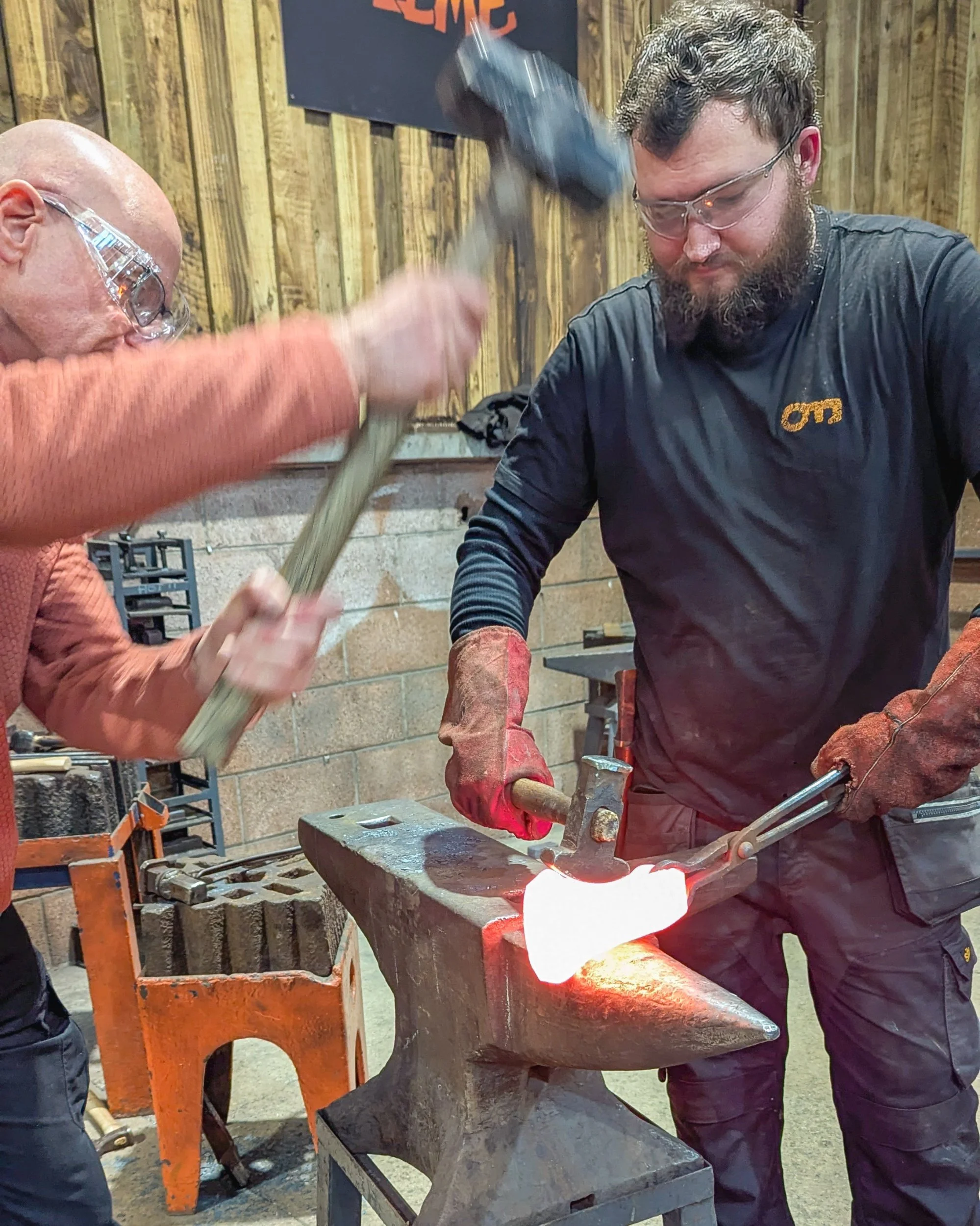 A close-up of a blacksmith at Oldfield Forge using a hammer and a specialized curved head tool to refine the side profile of a glowing orange axe head on the anvil.