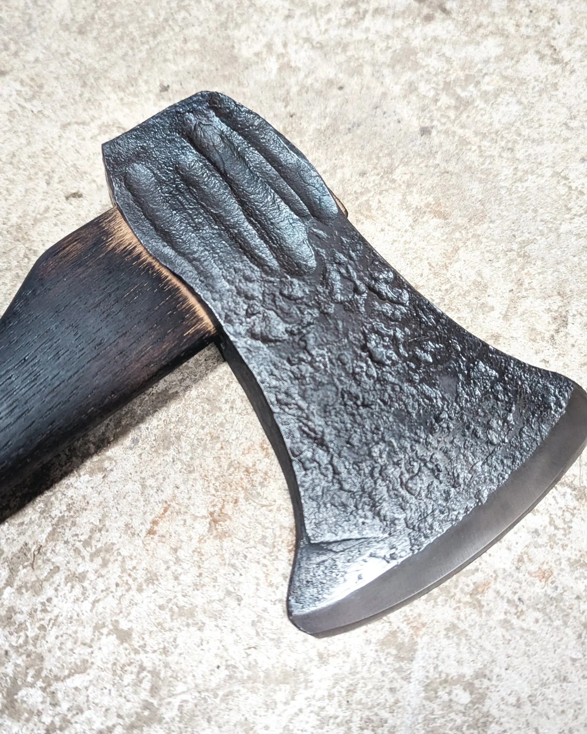 A macro close-up of a hand-forged axe head, showing the textural hammer marks on the cheek and the high-polish finish on the tempered cutting edge.
