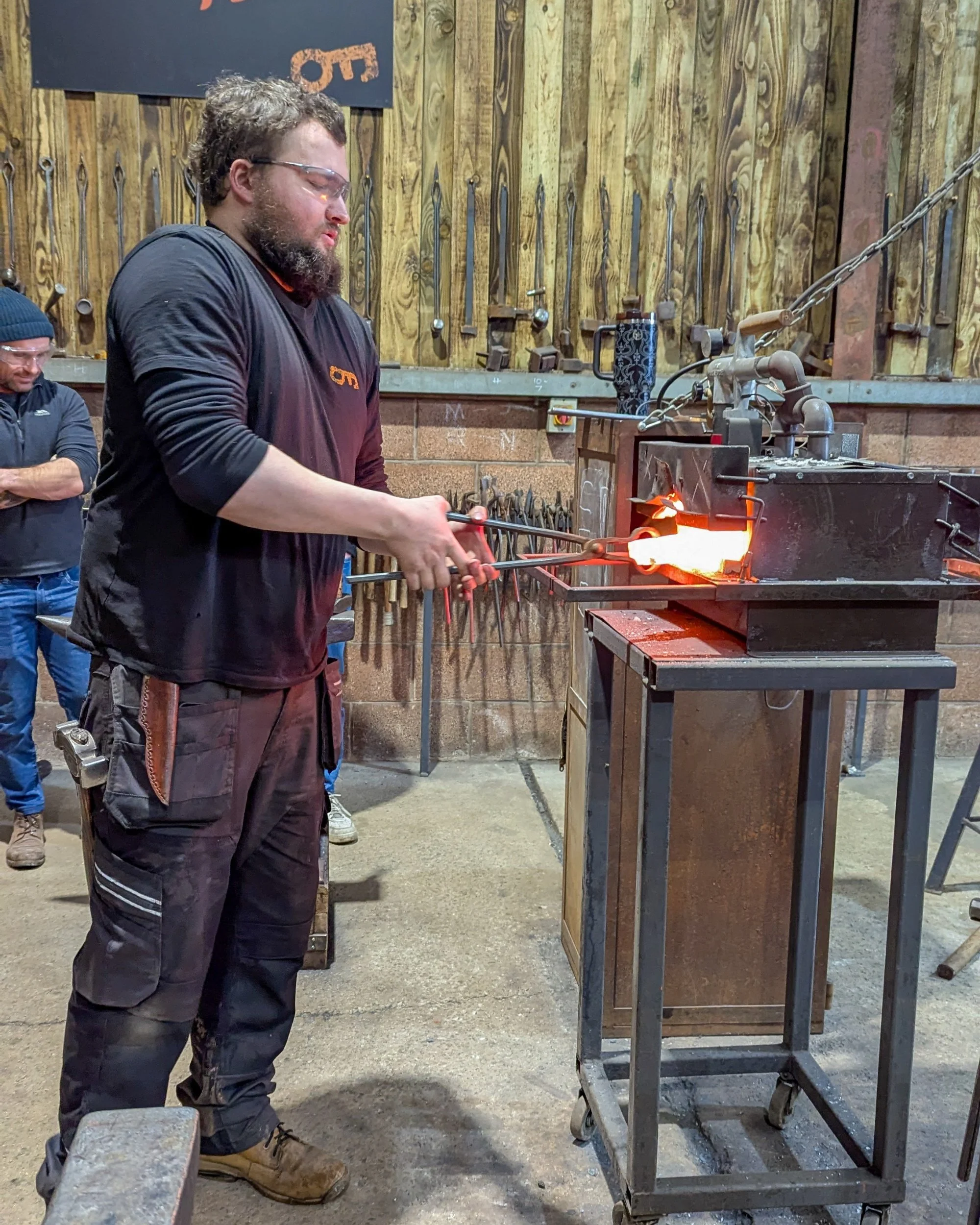 A blacksmith instructor using heavy-duty tongs to carefully remove a glowing, red-hot piece of metal from a traditional gas forge oven.