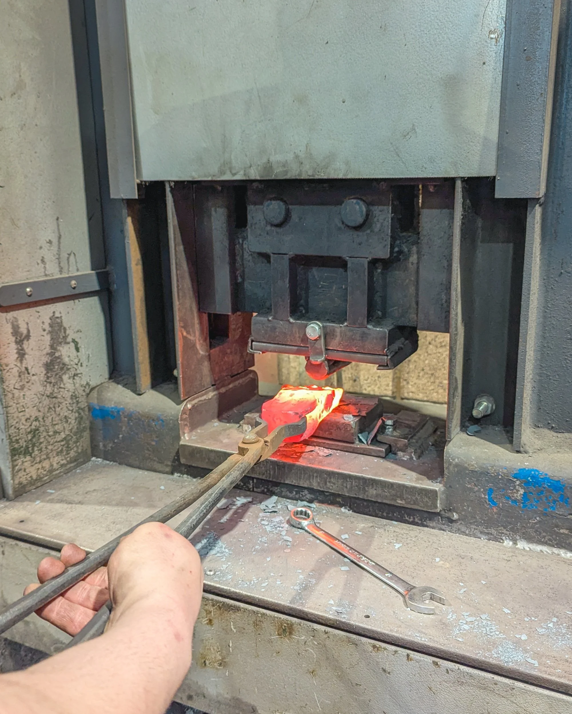 A close-up of a powerful hydraulic press at Oldfield Forge utilizing a specialized rounded fuller tool to indent and flow a glowing yellow-hot steel billet, applying extreme pressure to shape the metal.