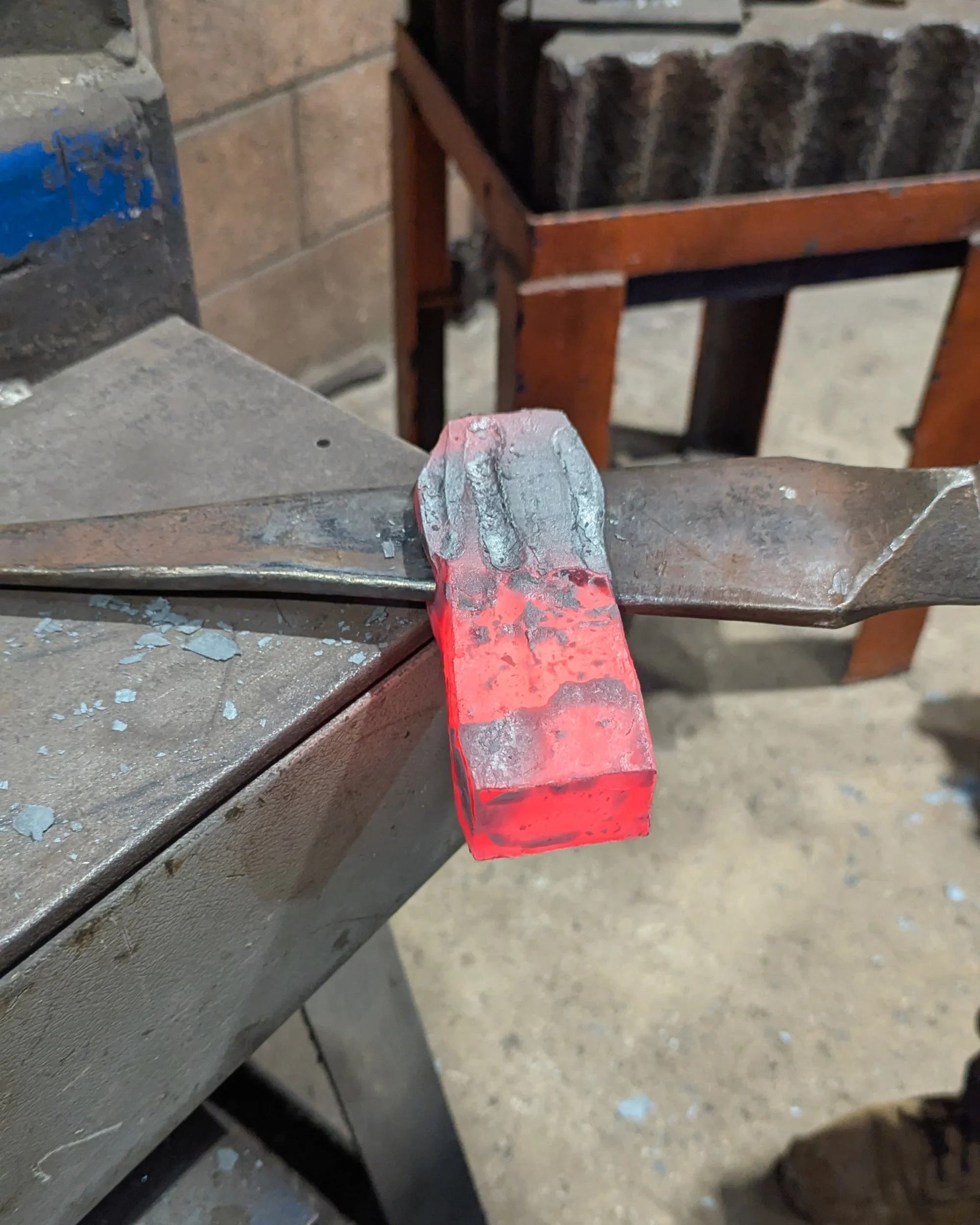 A close-up of a forged axe head with a steel drift still inserted in the eye; the metal shows a sharp contrast between the glowing orange heated section and the dark, cooled end.