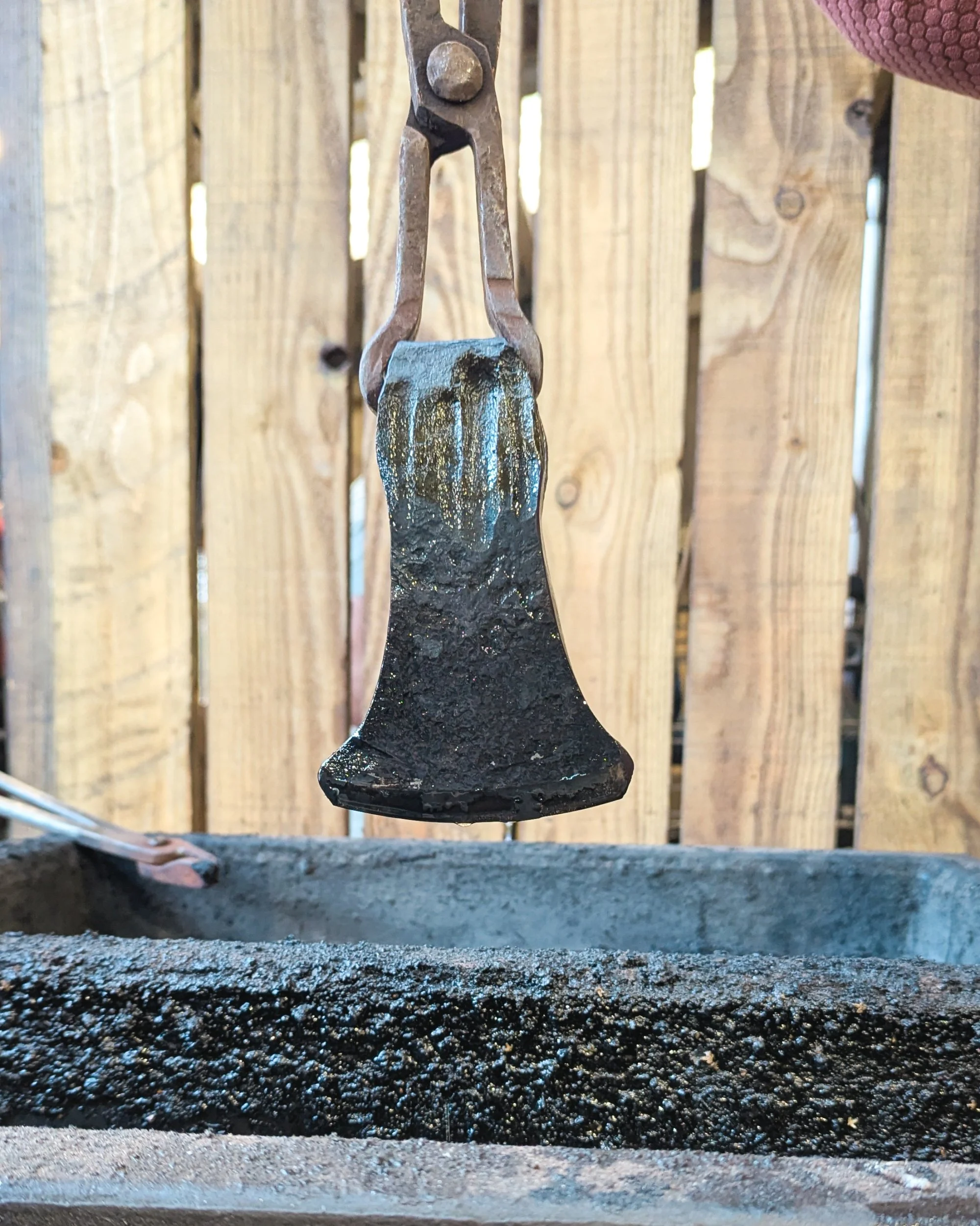 A blacksmith using long-handled tongs to hold up a forged axe head immediately after dunking it in a vegetable oil quench, with residual oil glistening on the dark, hardened steel.