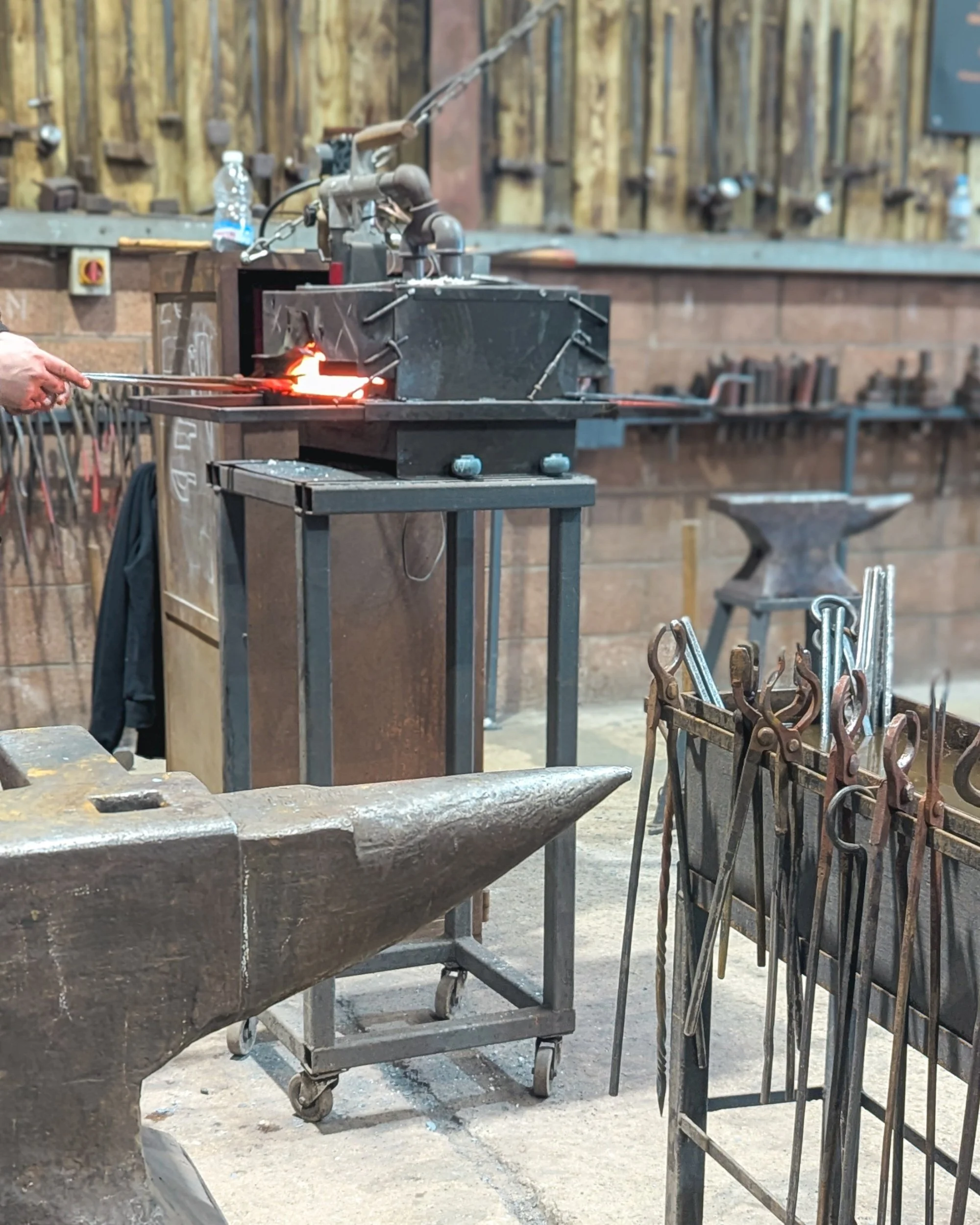 An overview of a structured Oldfield Forge workshop station, featuring a traditional anvil, a water quenching trough, and various tongs, with a blacksmith removing glowing metal from a forge in the background.