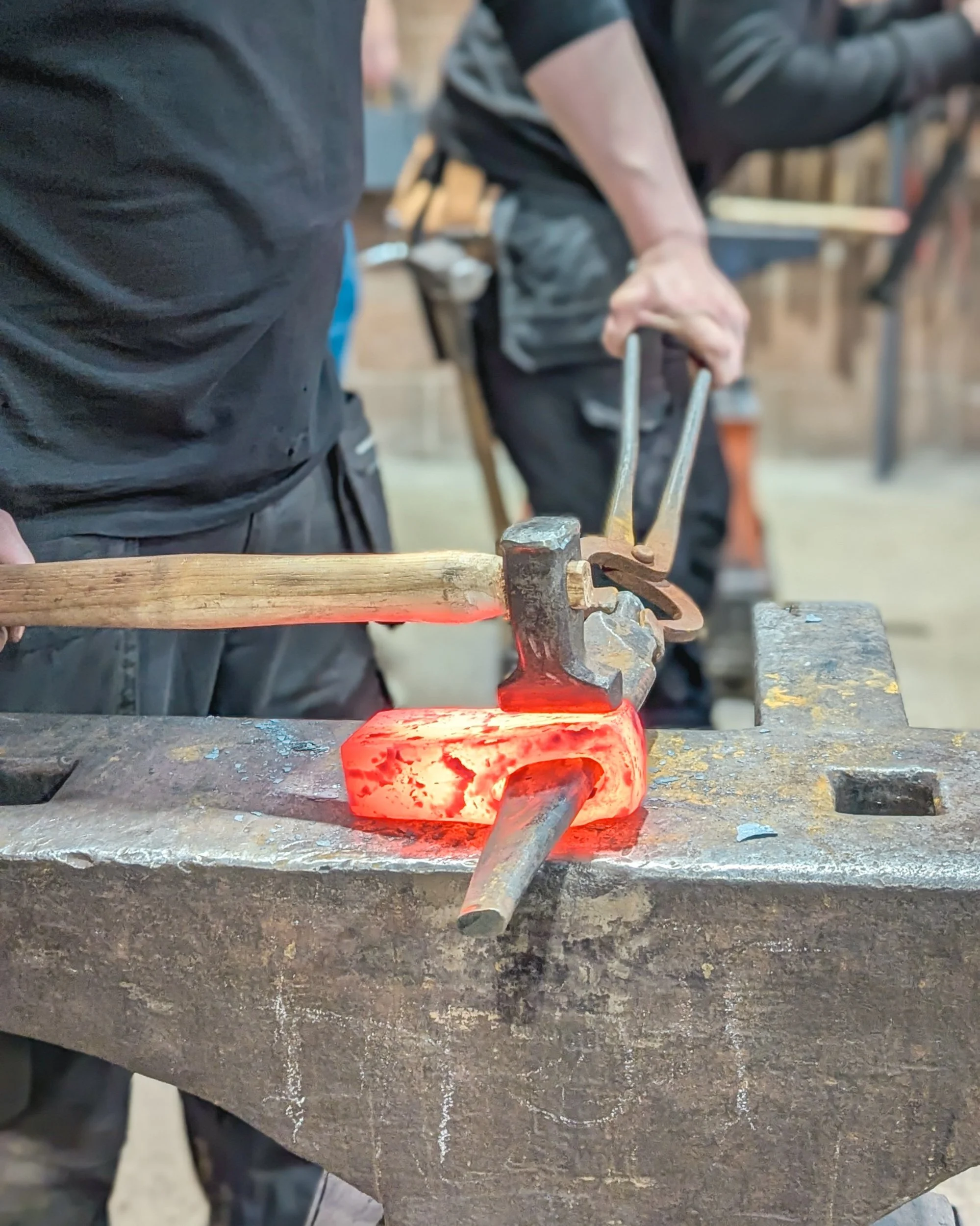 A blacksmith using heavy hammer strikes on the top flat of a glowing metal billet to begin shaping the distinct profile of an axe head on the anvil.