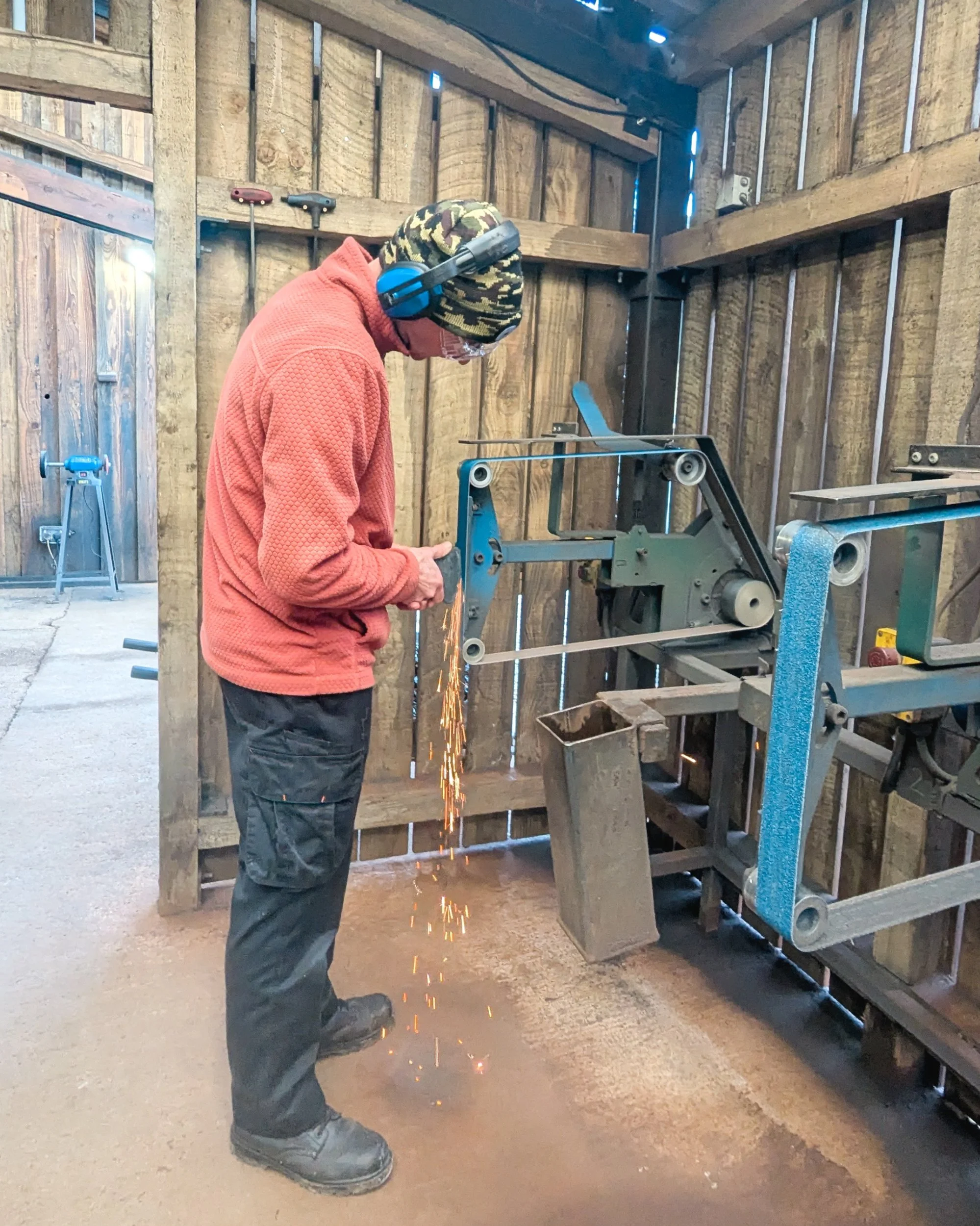 Using a belt sander to neaten the shape of a forged axe head, with sparks flying from the metal impact.