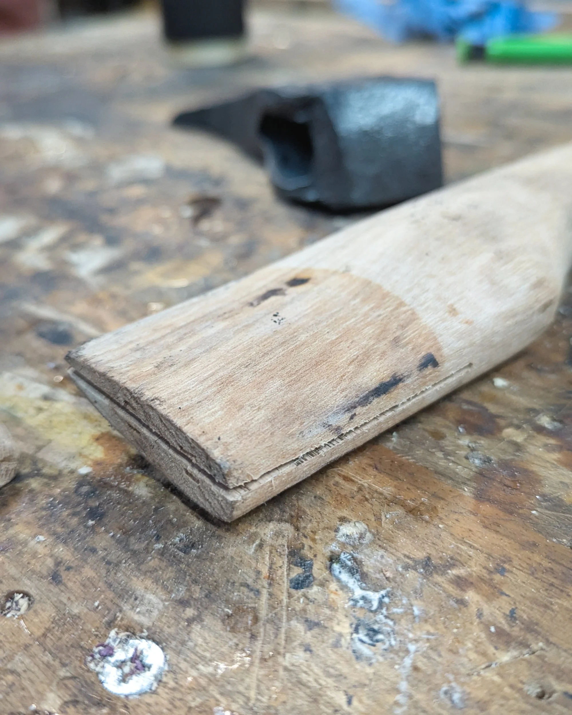 A macro close-up of the precision split cut into the top of a wooden axe handle, with the forged axe head blurred in the background awaiting assembly.