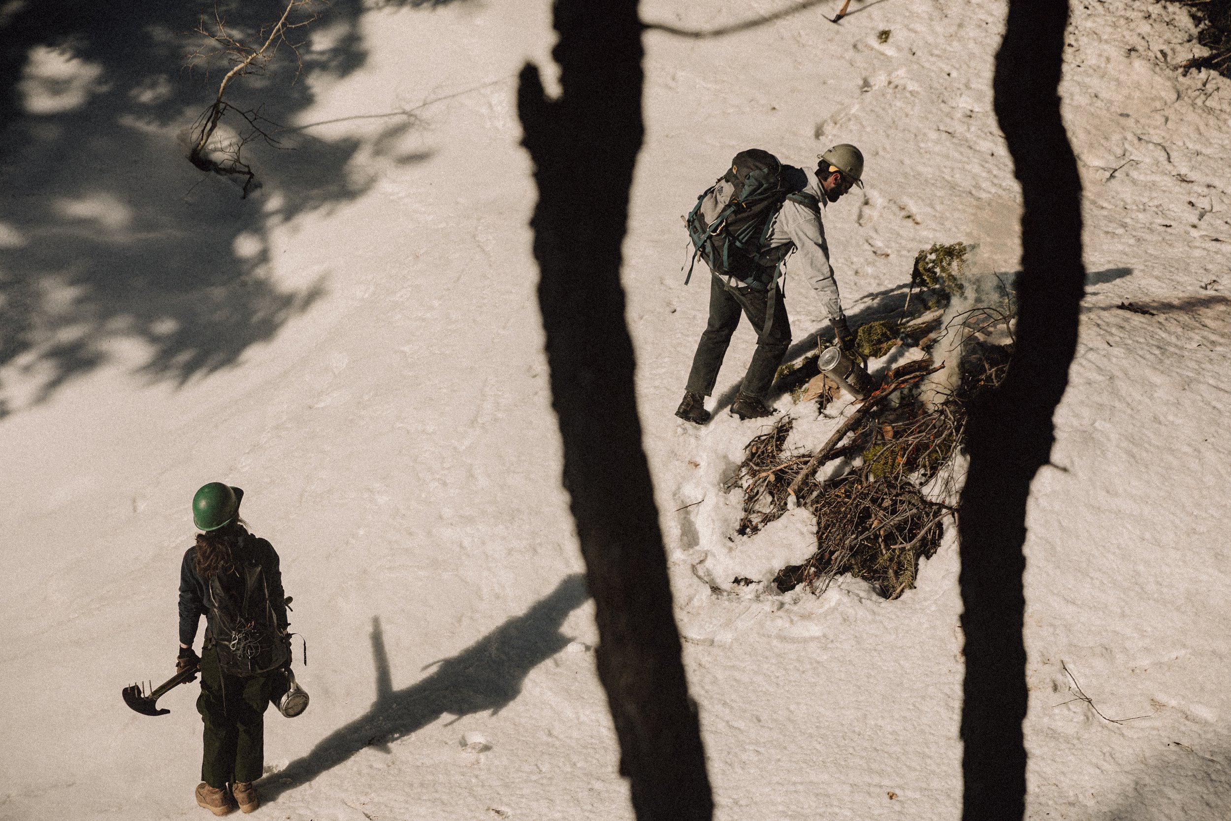 two people below walking across the snow under trees. one man is lighting a burn pile covered in snow, with a woman waiting holding a drip toeing and shovel as they wear backpacks