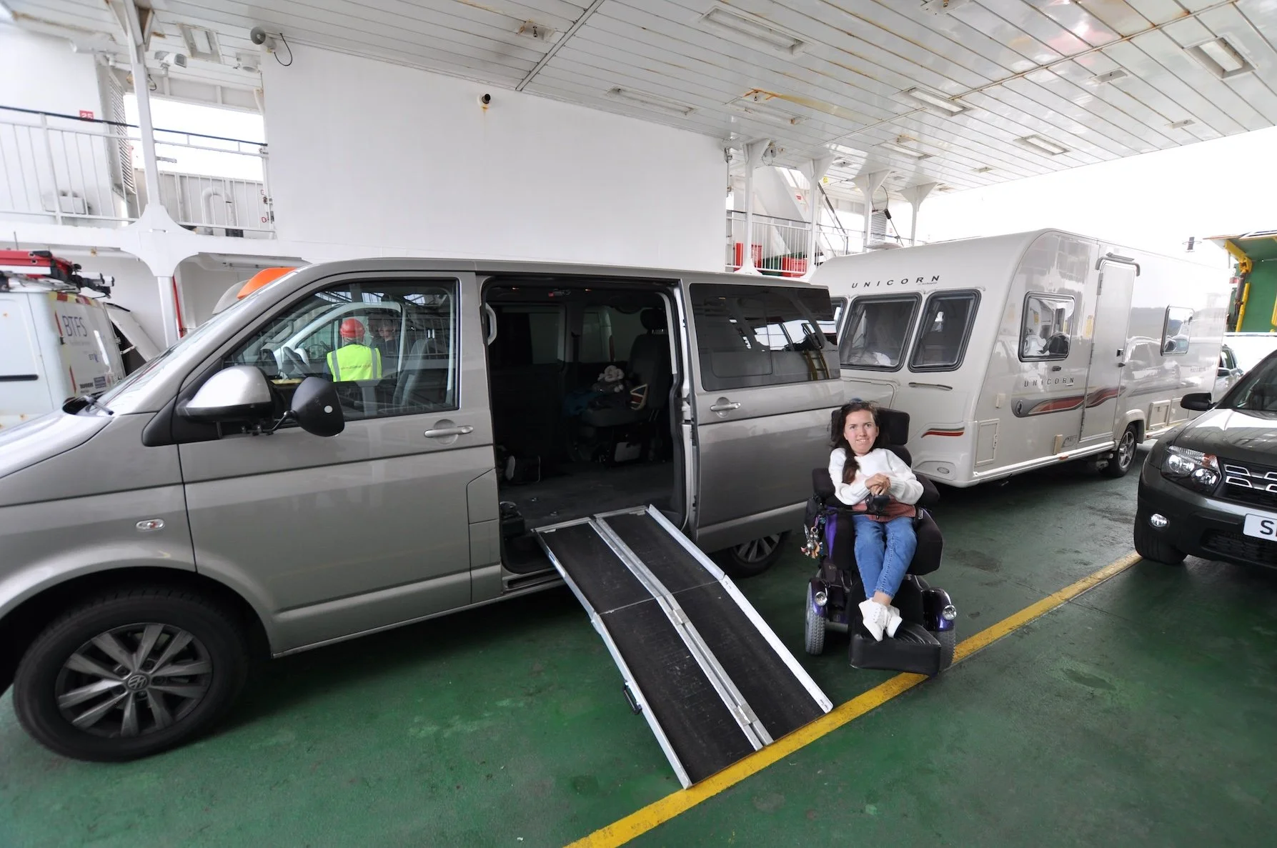 Karla in her Balder powerchair sitting next to her wheelchair accessible Volkswagen Transporter hitched up to a caravan, with a portable ramp leading up to the cars side door