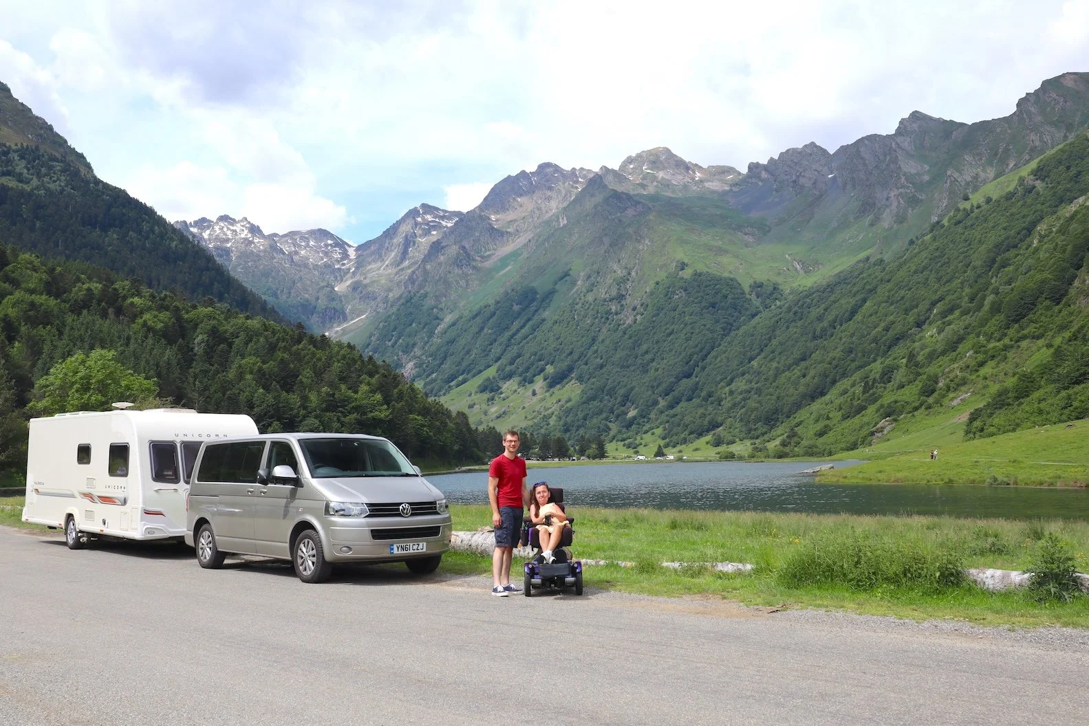 Stephen and Karla in the French Pyrenees standing next to their wheelchair accessible tow car and caravan, with a lake behing them surrounded by mountains.