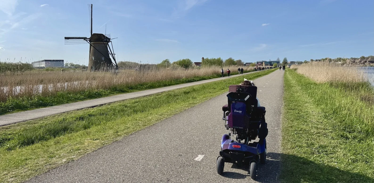Karla in her Balder powerchair, driving along a cycle path next to a traditional Dutch windmill at UNESCO Werelderfgoed Kinderdijk in the Netherlands