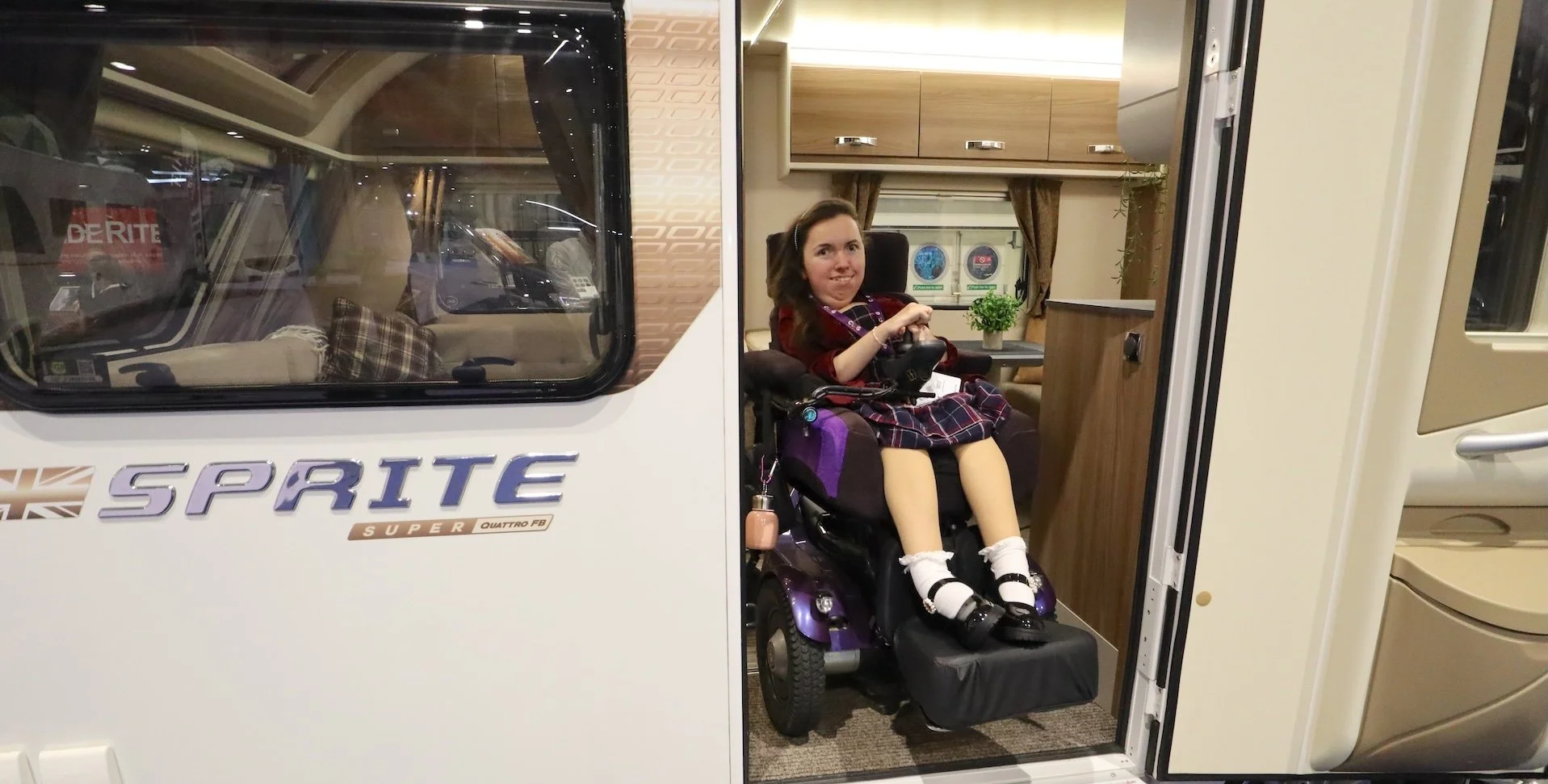 Karla, sitting in the doorway of a wheelchair accessible caravan in her Balder Powerchair. The caravan is a LIV Swift Sprite Super Quattro FB, modified by Coachbuilt.