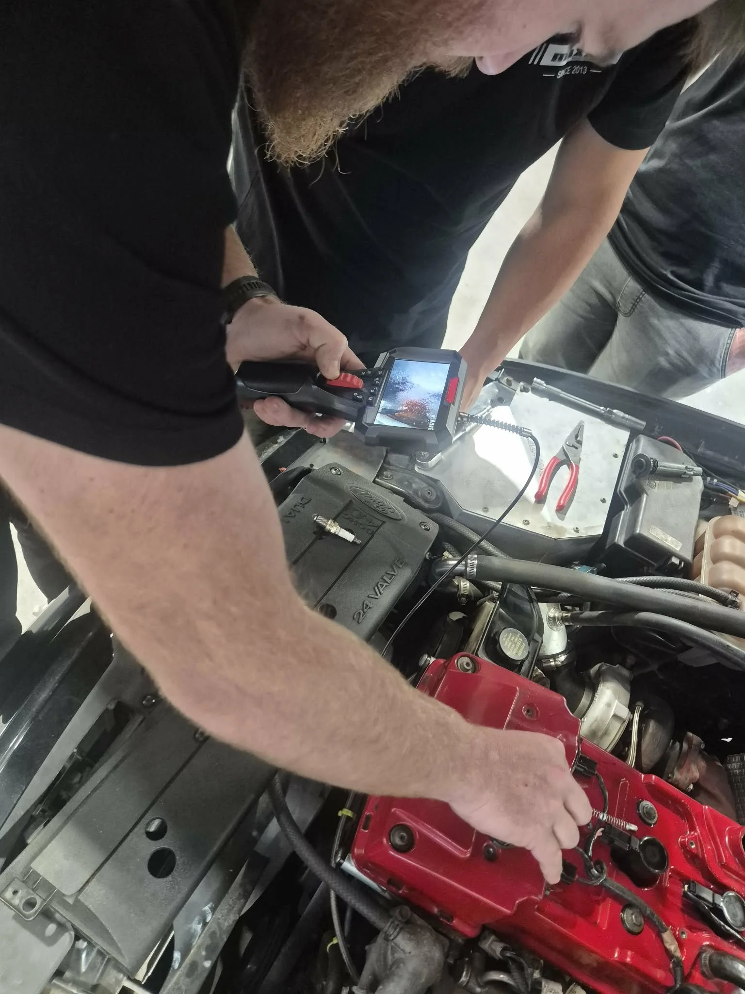 Close-up of a person using a diagnostic tool on a red car engine, with another person observing.