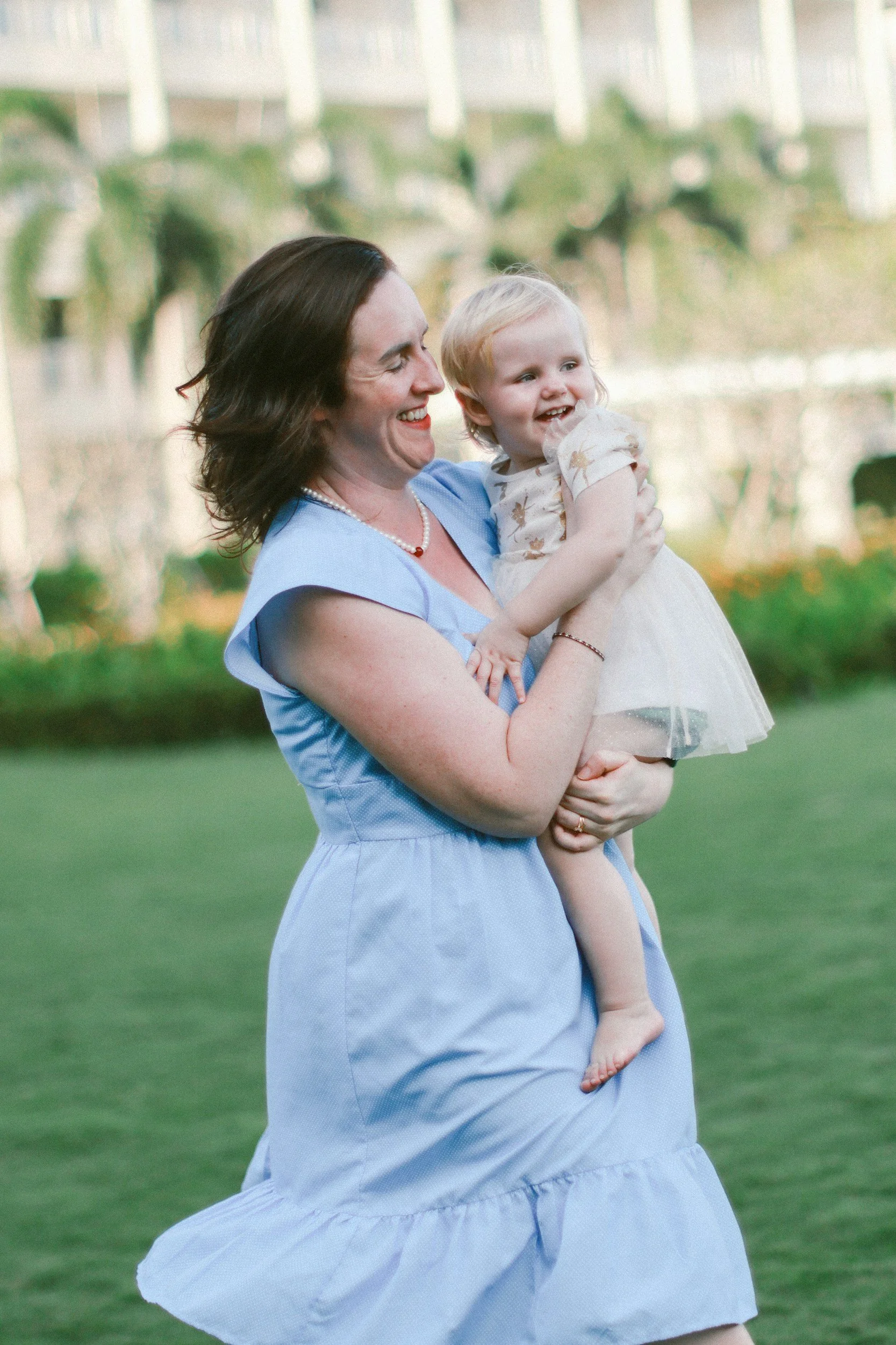 A woman in a blue dress holds a smiling toddler in a party dress. They are in a garden.
