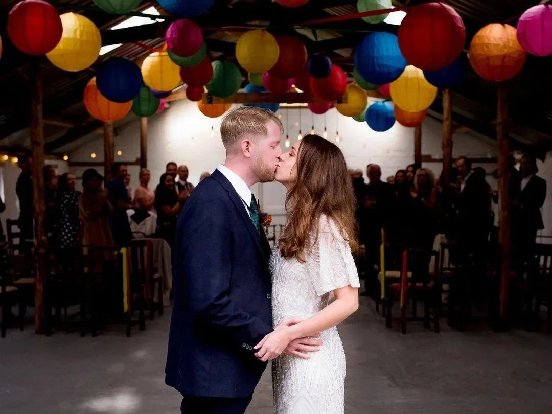 A young couple kiss on their wedding day, against a backdrop of colourful lanterns and their clapping wedding guests. They are having a Humanist wedding ceremony in a trendy barn.