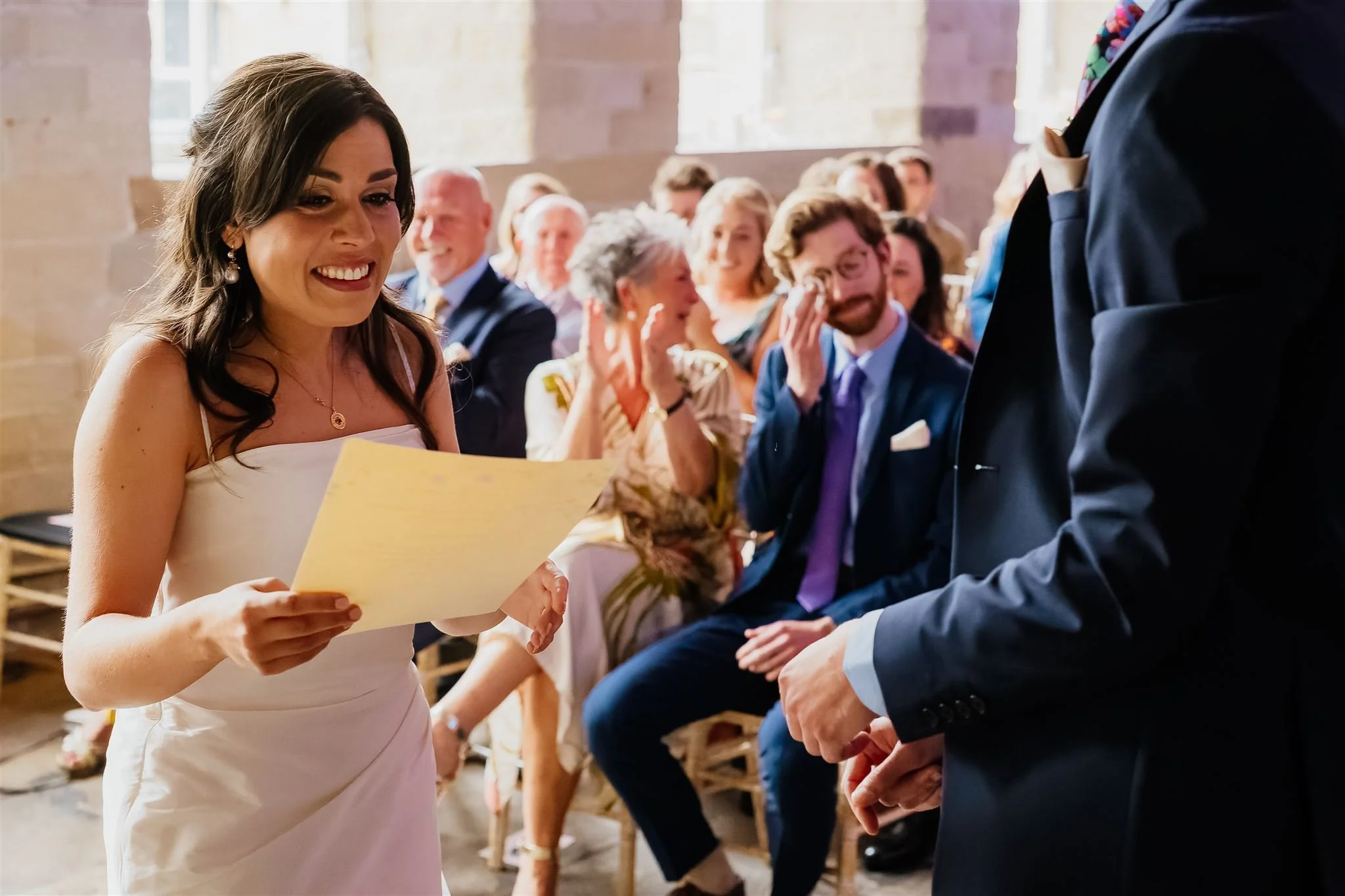 A smiling bride with dark hear in a white wedding dress reads her wedding vows from a sheet of paper. Her groom is in the foreground, we cannot see his face. In the background wedding guests display a range of emotions - smiling, clapping and crying