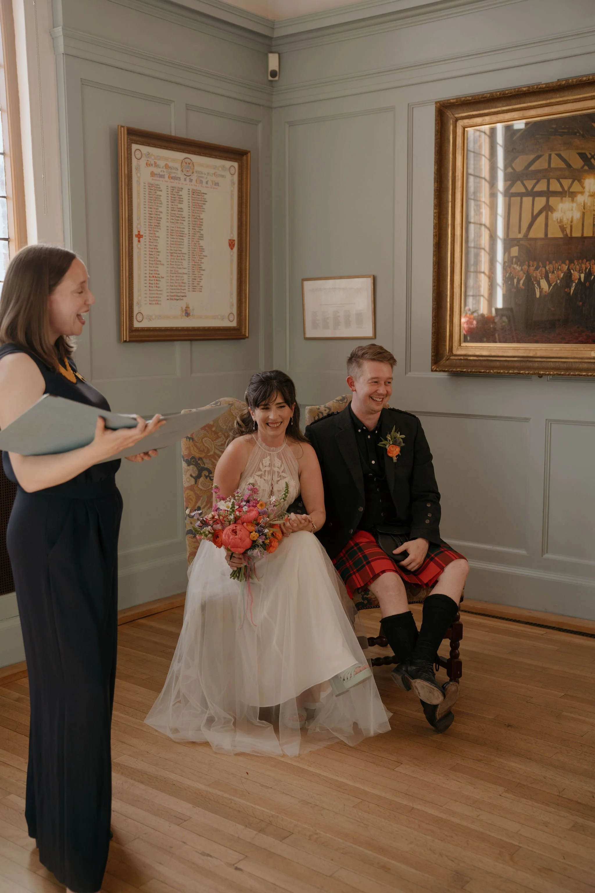 A female wedding celebrant reads from a folder to a bride and groom. The groom wears a red tartan kilt and the bride a white halterneck dress; they are both smiling broadly during their wedding ceremony.