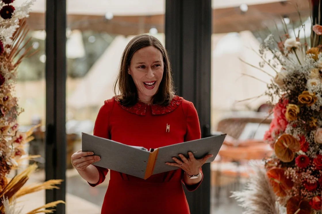 Rachael, a smiling white woman with brown hair, reads from a blue folder during a Humanist ceremony. She looks like she's enjoying herself!