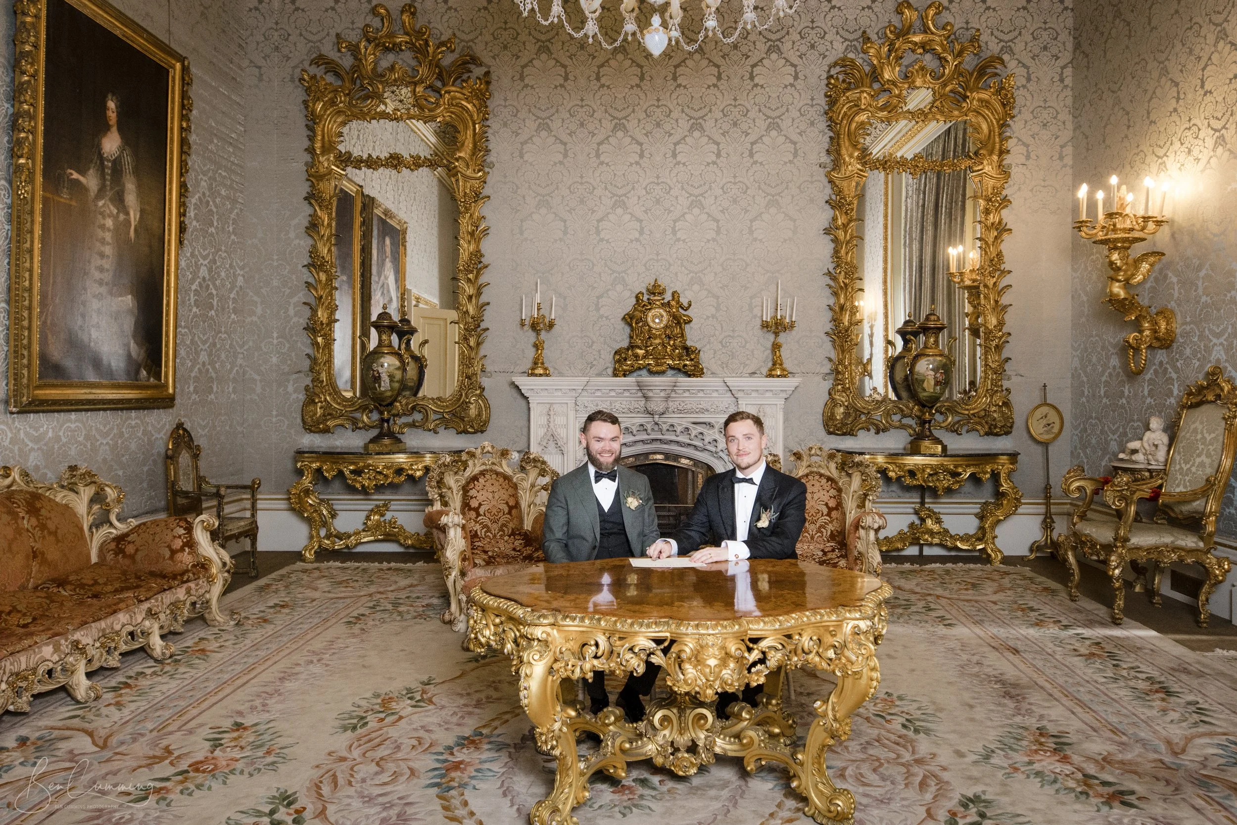 Two smartly dressed men sit at an ornate gold table on their wedding day, surrounded by luxurious gilt furniture in an English stately home.