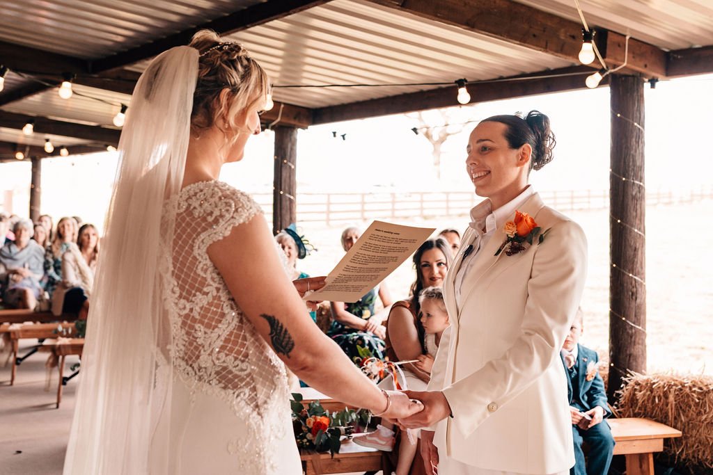 Two LGBTQ+ women hold hands as they make their wedding vows to each other. One wears a white dress and veil and the other wears a white suit. In the background are smiling wedding guests.