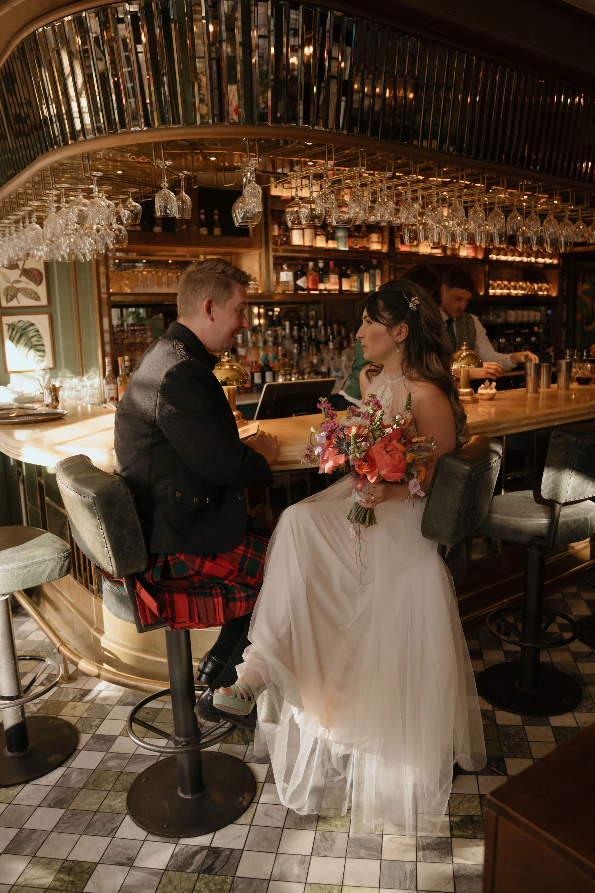 A bride and groom sit at a stylish bar. He wears a red kilt and she is carrying a bouquet of flowers and wearing a white wedding dress.