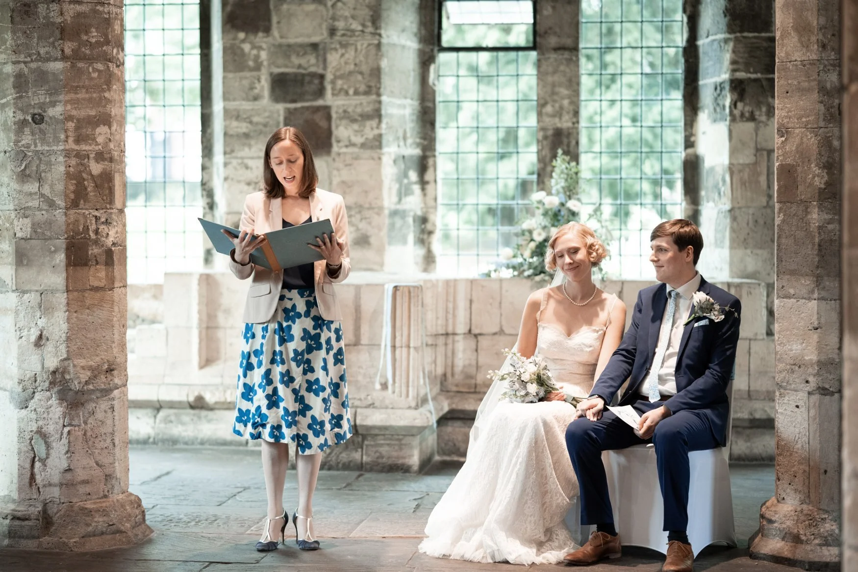 Leading Yorkshire wedding celebrant reads from a blue folder in a Medieval stone building. A smiling bride and groom sit beside her during their wedding ceremony.