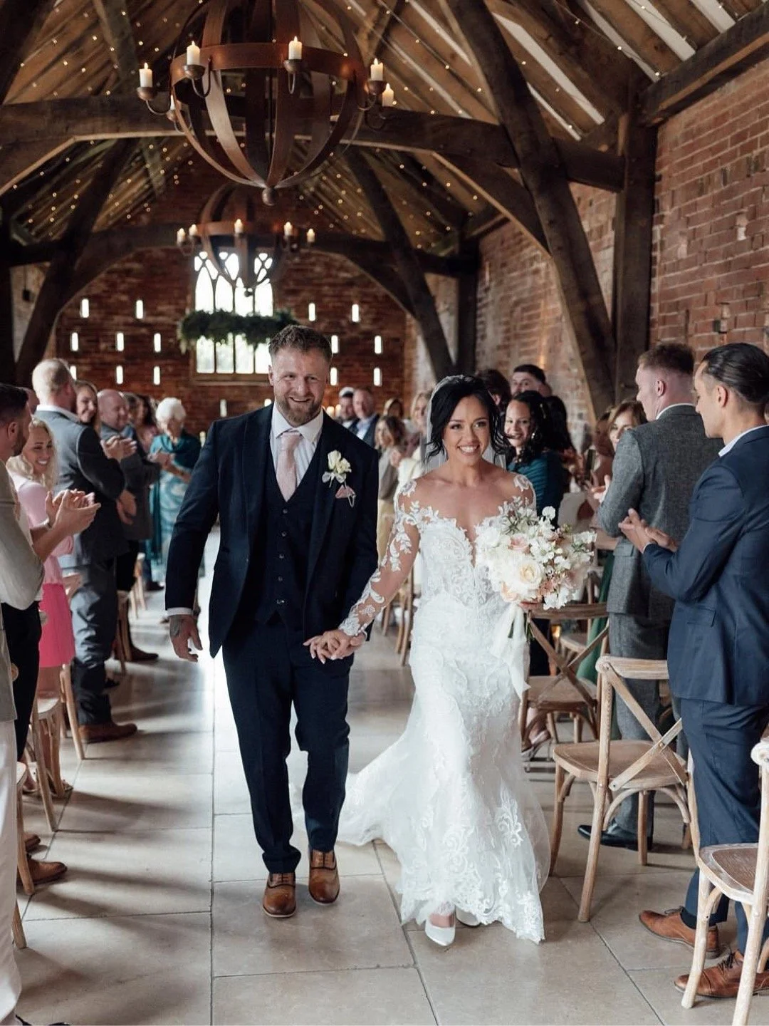 A bride and groom walk down the aisle after their humanist wedding ceremony. They are both smiling, and their wedding guests are clapping.