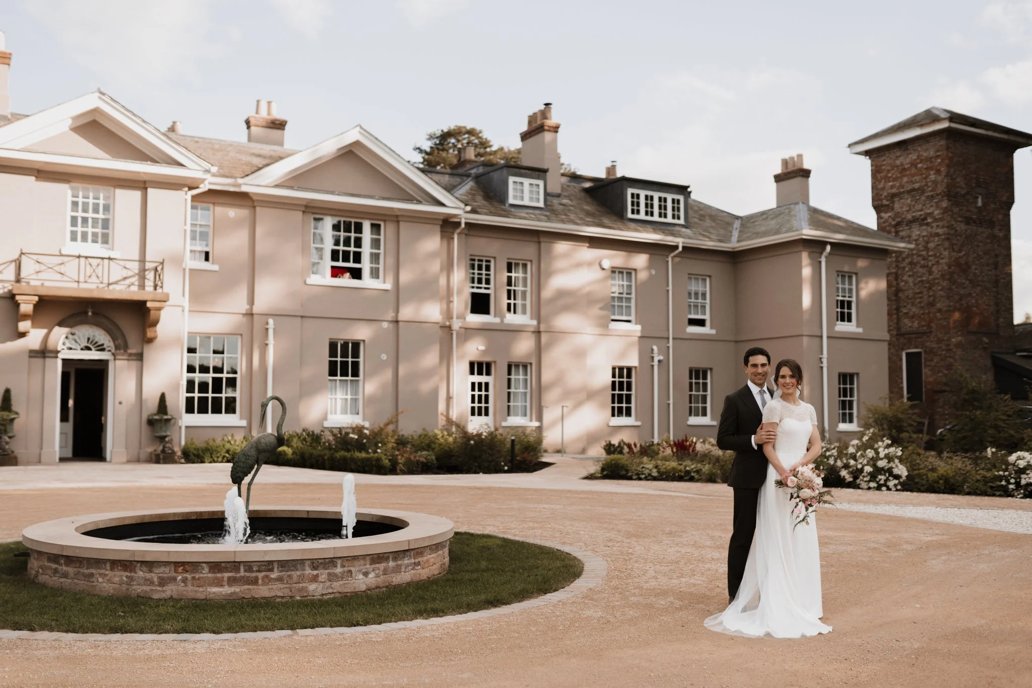 A bride and groom stand in front of an Italianate mansion next to a fountain on a sunny day.