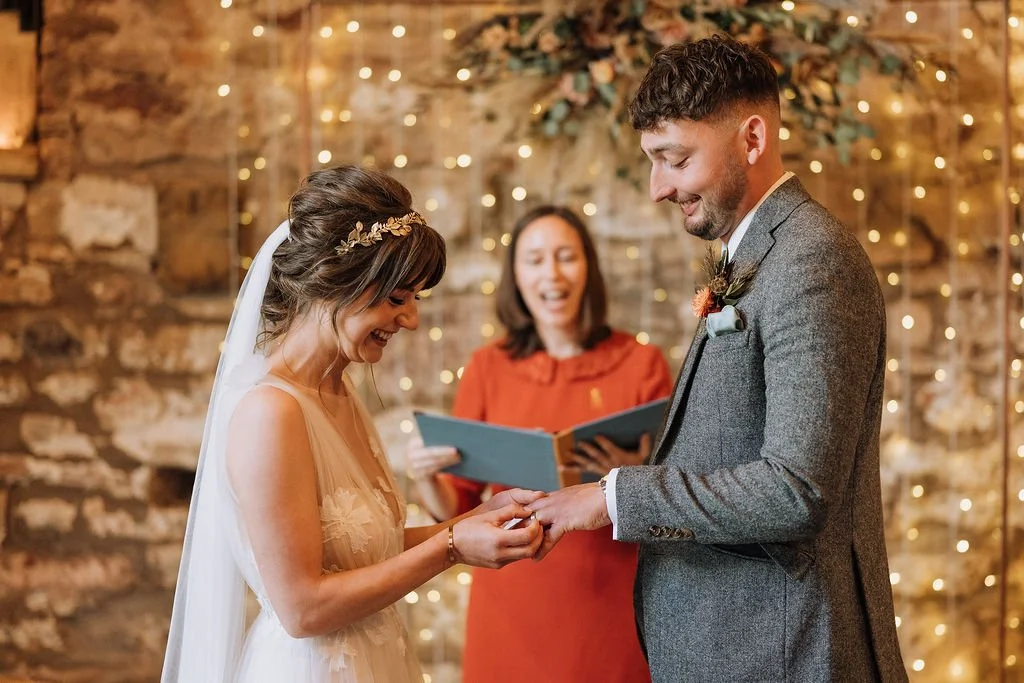 In a rustic barn lit with fairy lights, a smiling celebrant watches on as a bride slides a wedding ring onto her groom's finger.