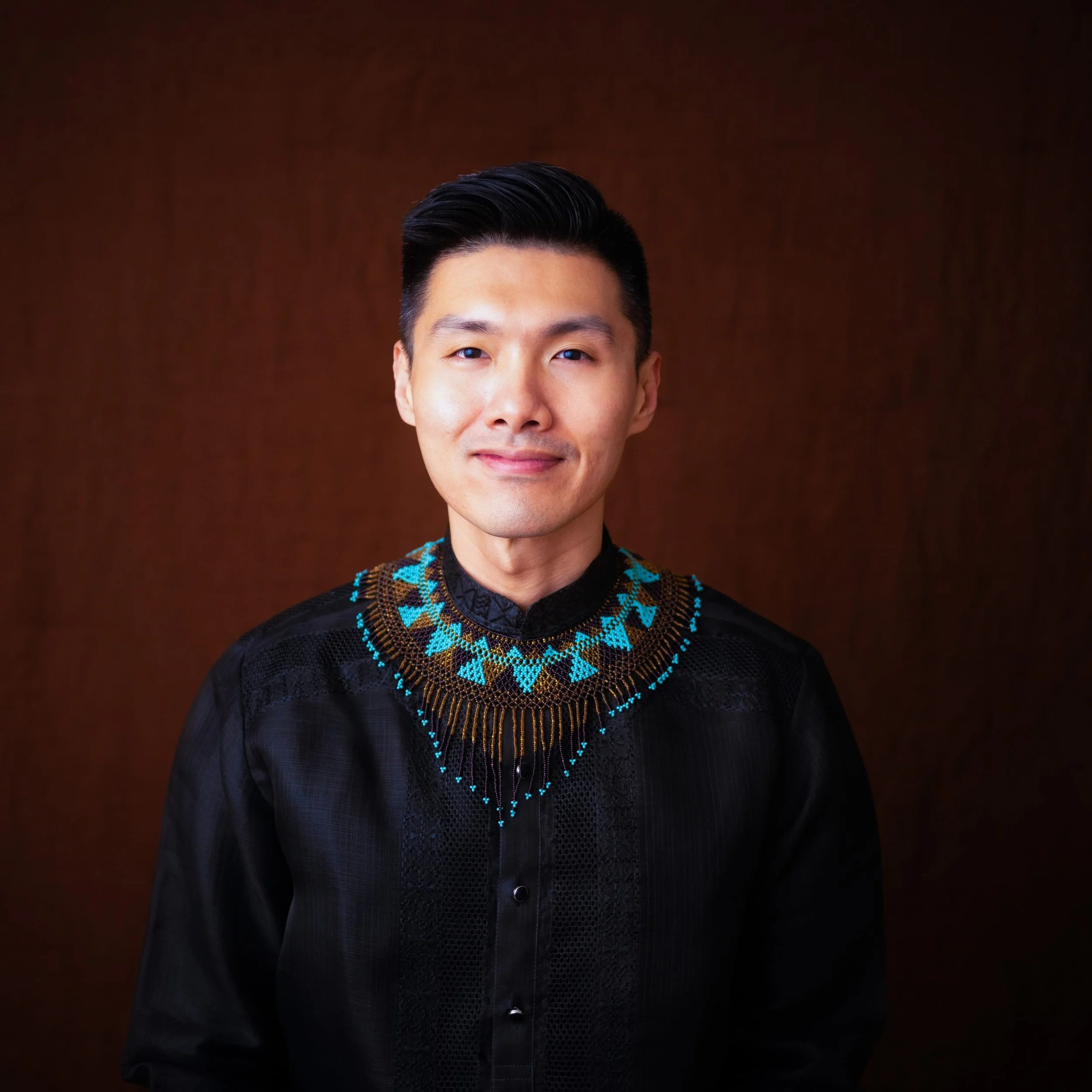 Close-up of a man with black hair, wearing a black shirt with an intricate, colorful beaded necklace pattern.