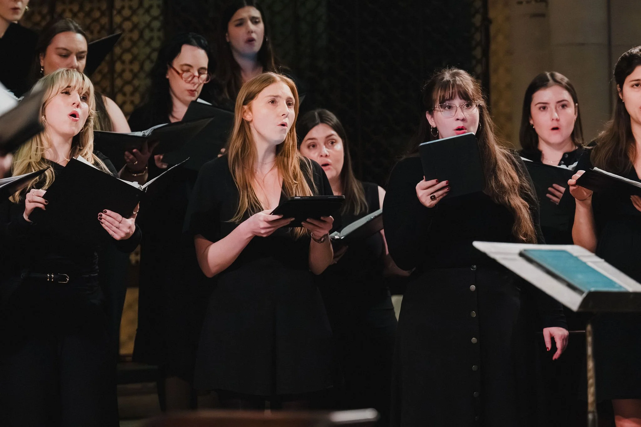 A group of singers performing, holding black songbooks, with some singing and looking at the conductor's sheet music.