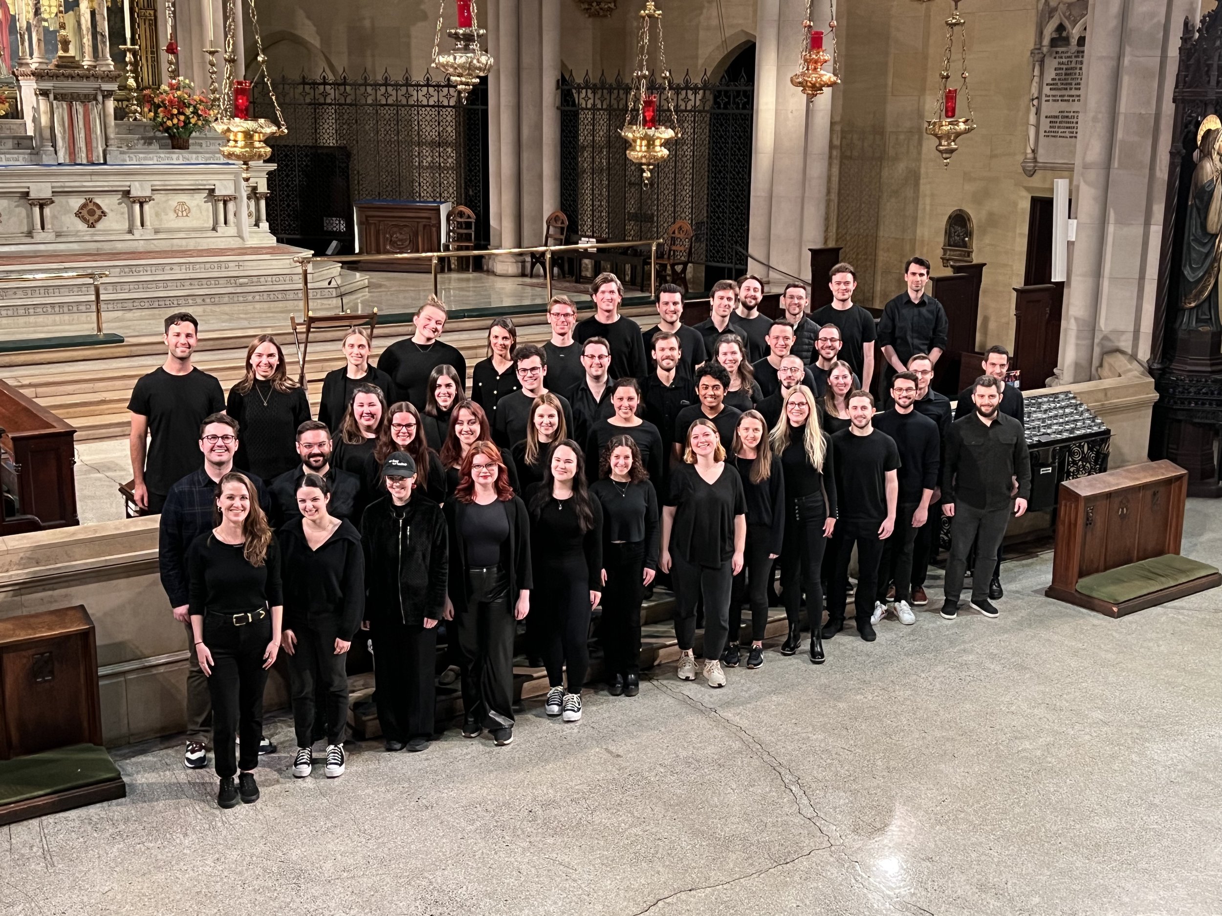 Group of people standing in a church, smiling for a group photo. The church features an altar with floral arrangements and religious statues, with candles hanging from the ceiling.