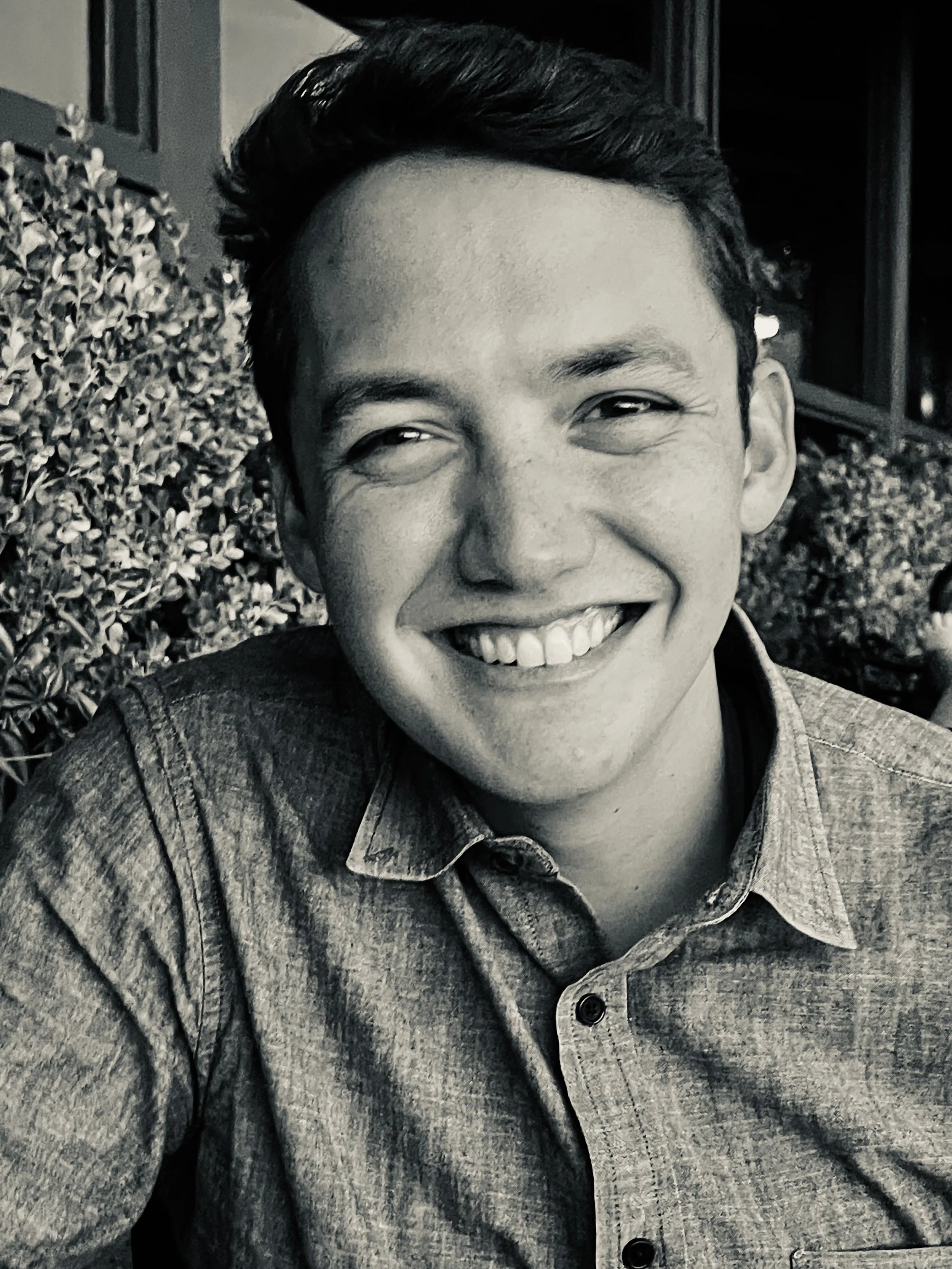 A young man smiling, wearing a denim shirt, with short dark hair, in an outdoor setting with bushes and a building in the background.