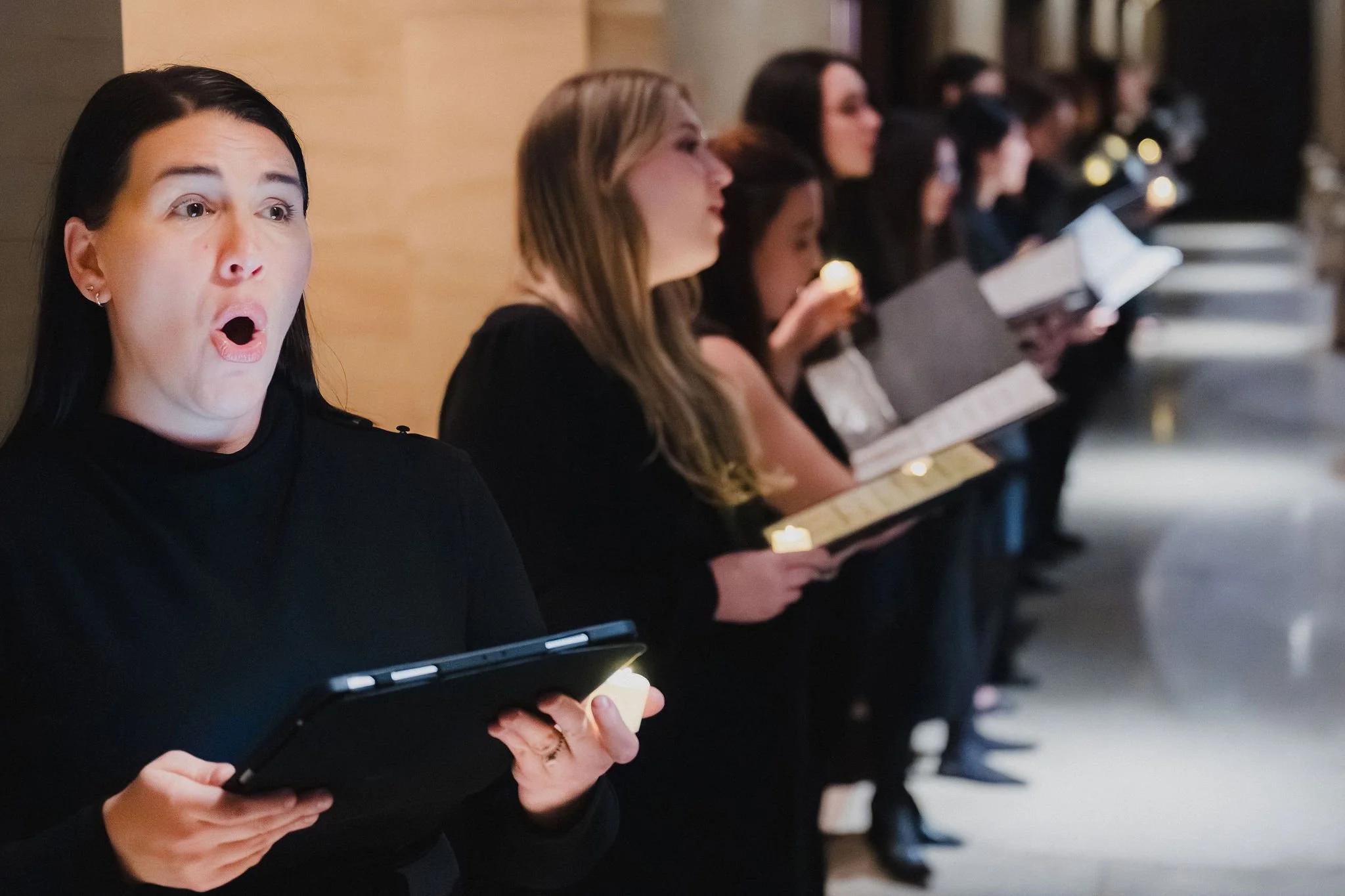 A group of singers dressed in black are standing in a line, holding candles, and reading from booklets during a religious or memorial service.