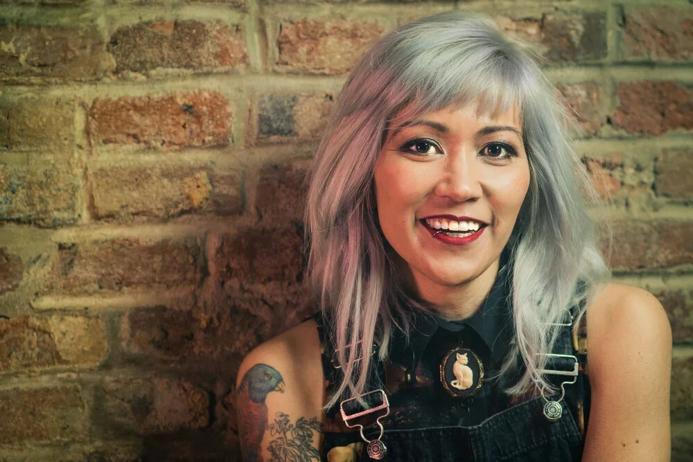 A woman with light gray, shoulder-length hair, smiling, wearing black overalls with a pin and a tattoo on her arm, standing against a brick wall.