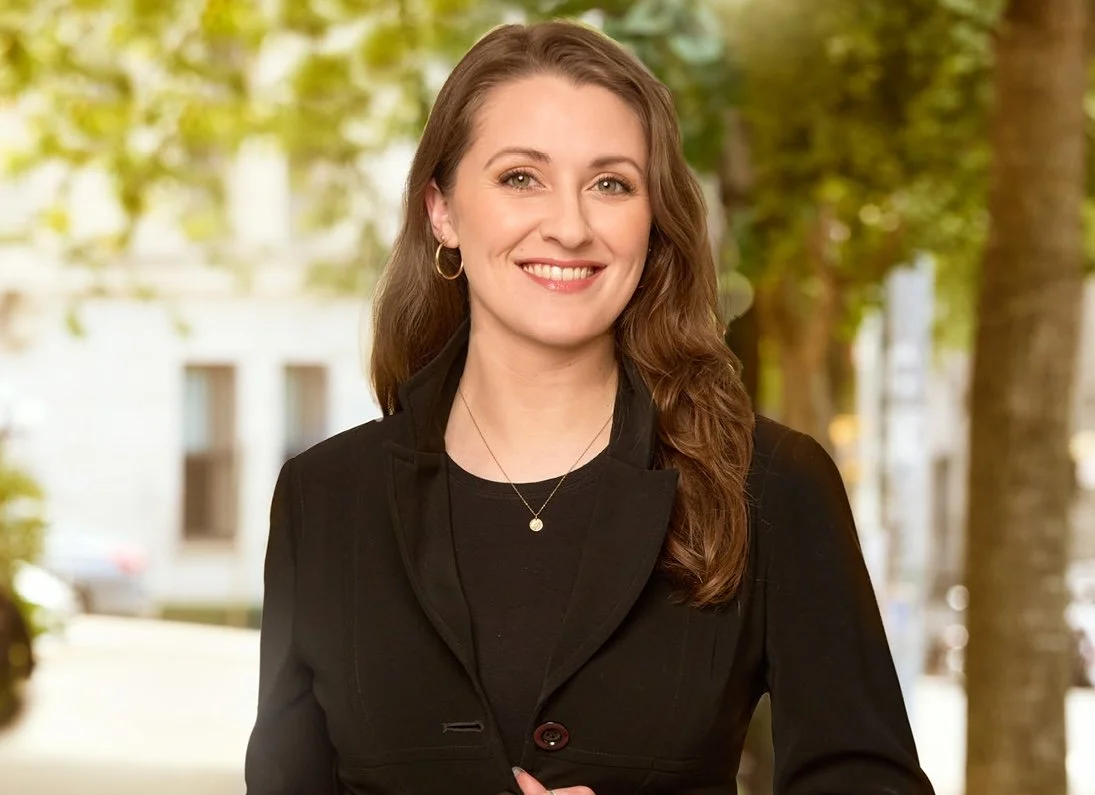 A woman with long brown hair in loose curls, wearing a black blazer and a black top, standing outdoors in front of trees and a city street, smiling at the camera.