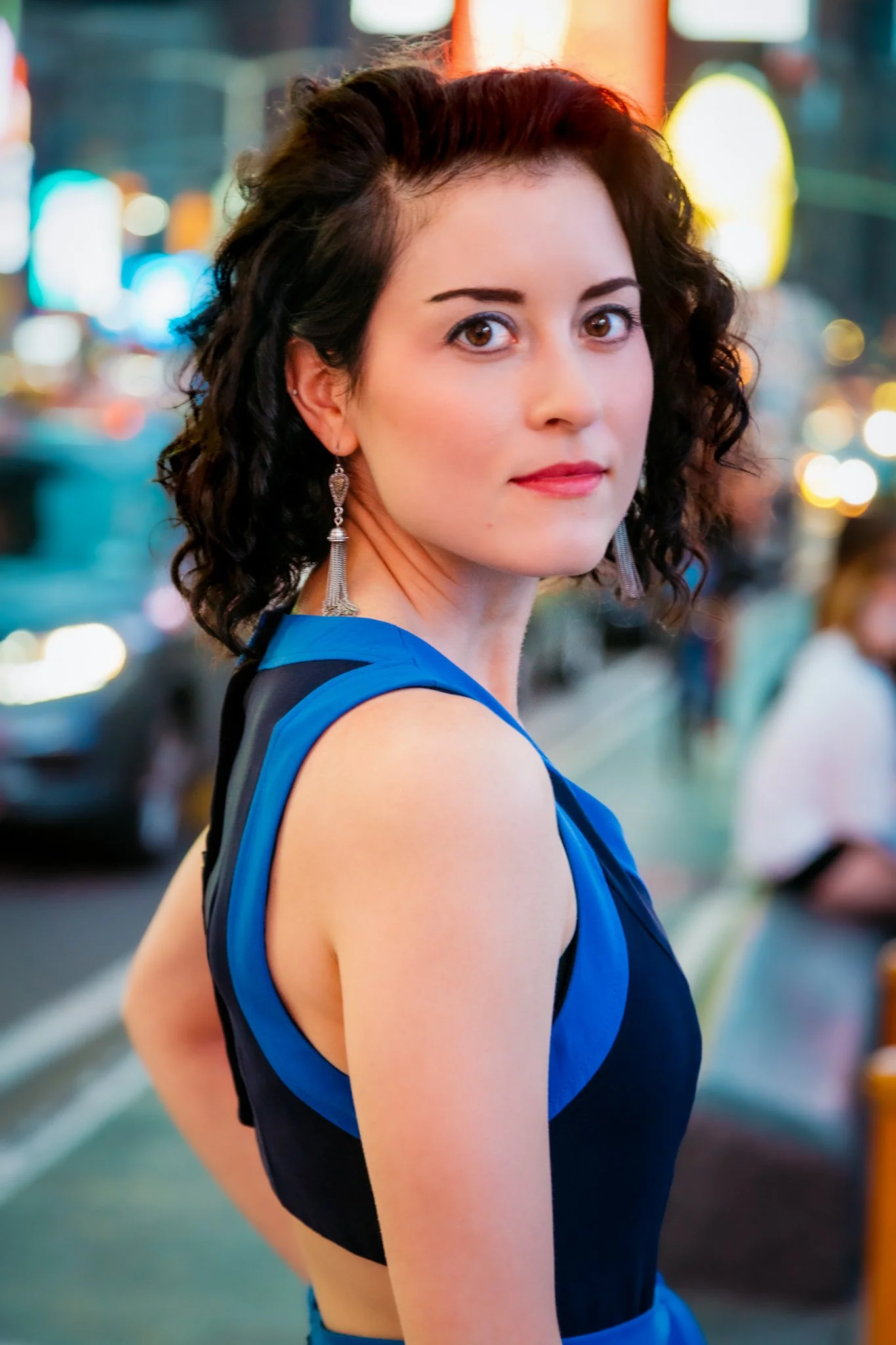 A woman with curly dark hair wearing earrings and a blue dress, standing on a city street at night with colorful blurred lights in the background.