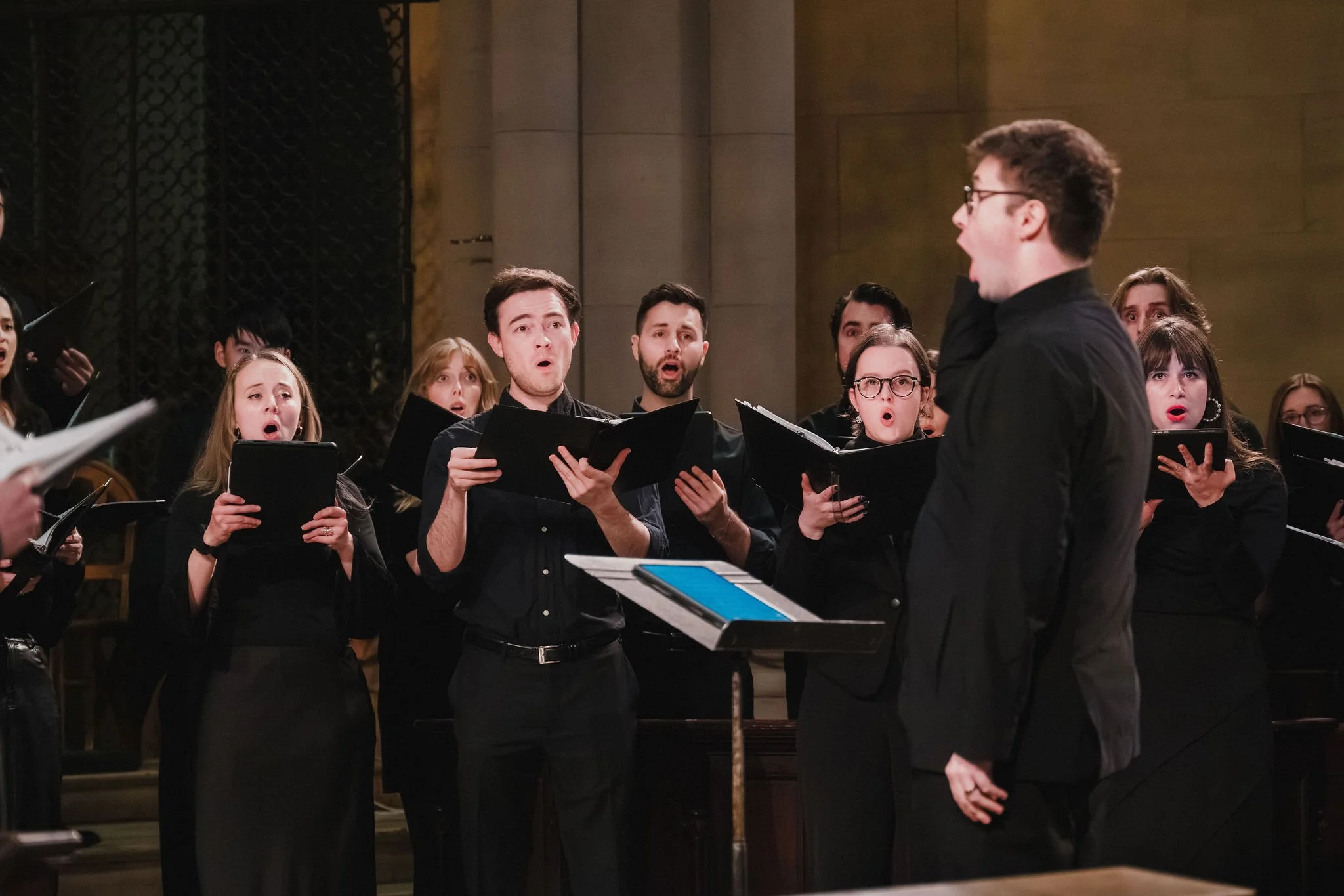 A group of choir singers singing, led by a conductor, in a concert or rehearsal setting.