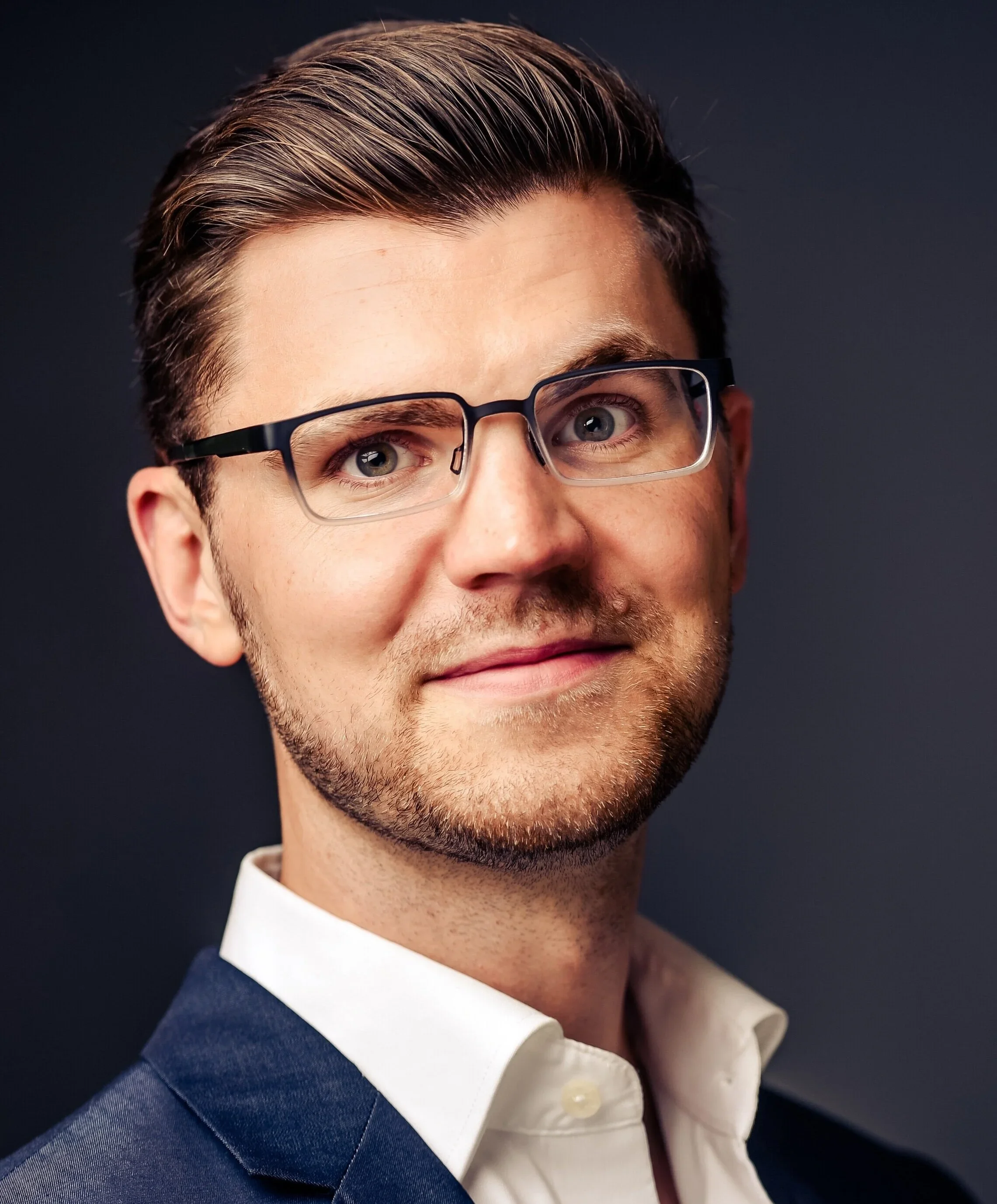 Close-up of a young man with neatly styled brown hair, wearing glasses with black frames, dressed in a white shirt and dark suit jacket, smiling slightly against a dark background.