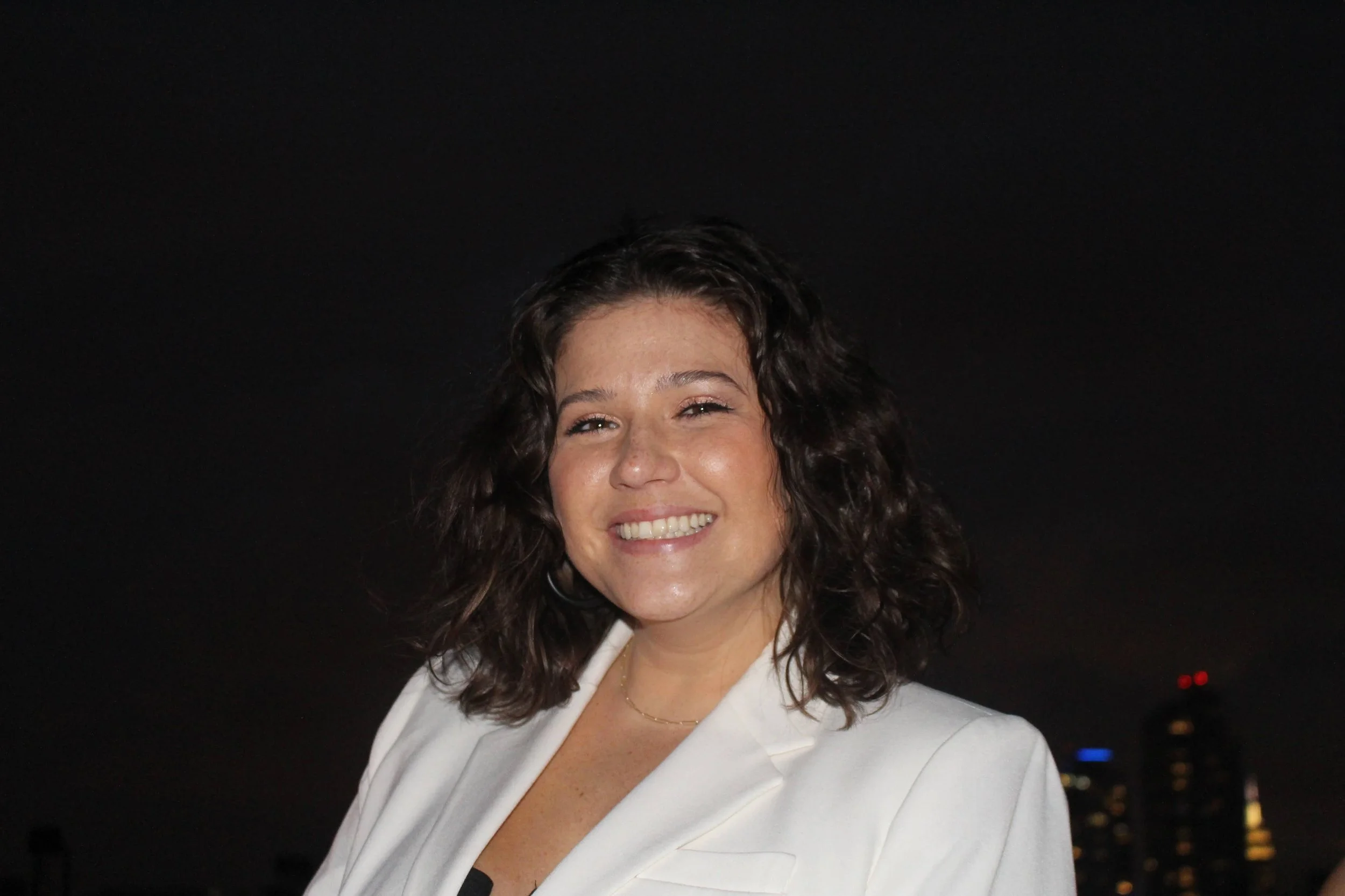 A smiling woman with dark curly hair wearing a white blazer, taken at night with city lights in the background.