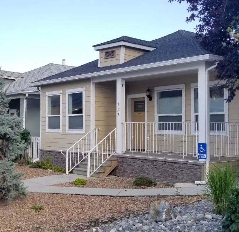 A beige house with white trim and a dark roof features a front porch with a wheelchair accessible ramp and steps. The house number 727 is visible, and there is a blue sign indicating handicapped parking behind the porch. The yard has decorative rocks, small bushes, and landscaping.