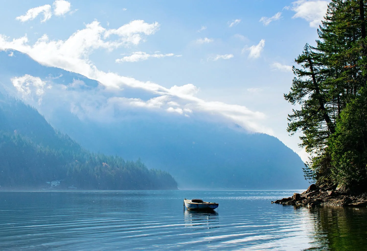 A boat floating on a calm lake with forested mountains and blue sky with clouds in the background.