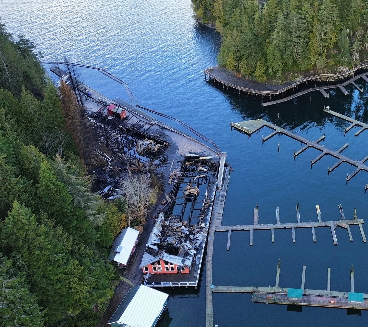 An aerial view of a damaged building by the water, with some parts collapsed and charred, adjacent to a forested area and a marina with empty docks.