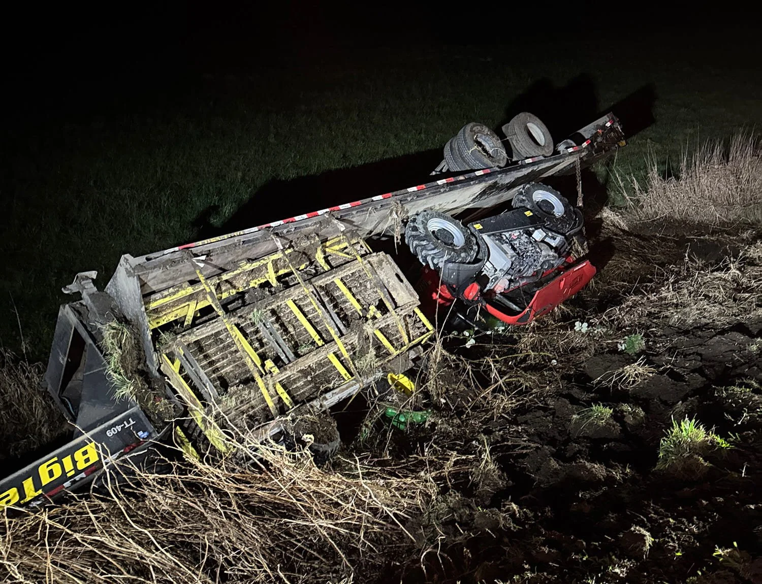 Upside-down truck with its undercarriage exposed, lying in a muddy area at night with grass around.