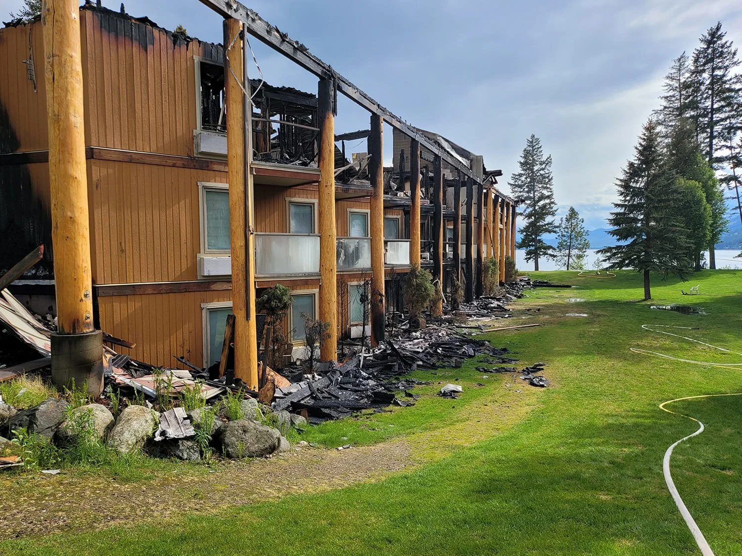 Burned multi-story building with damaged wooden pillars and debris scattered on the ground, near a grassy area with tall trees and a lake in the background.
