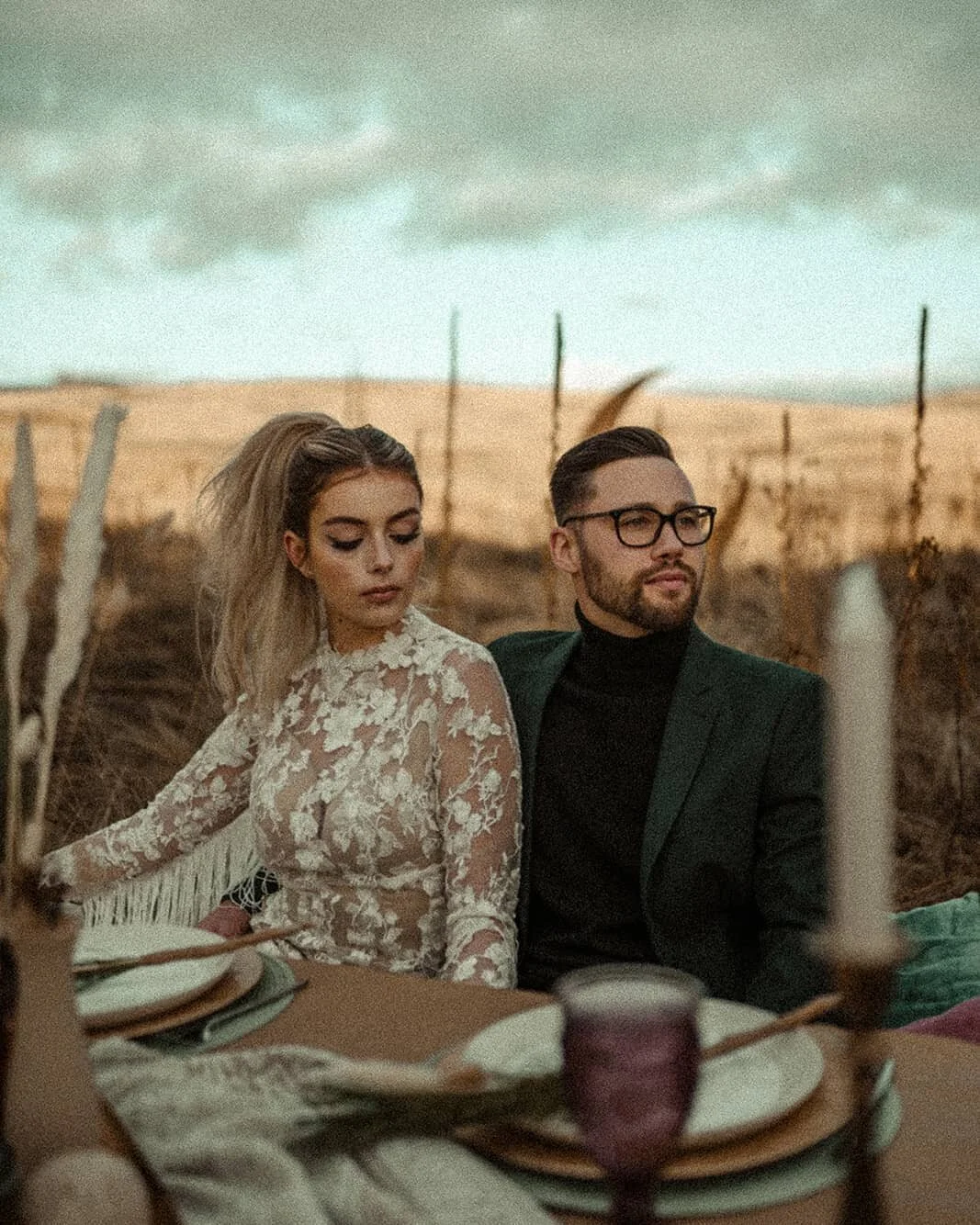 A man and woman sitting outdoors at a dining table with plates and glasses, in a rural field with a cloudy sky in the background.