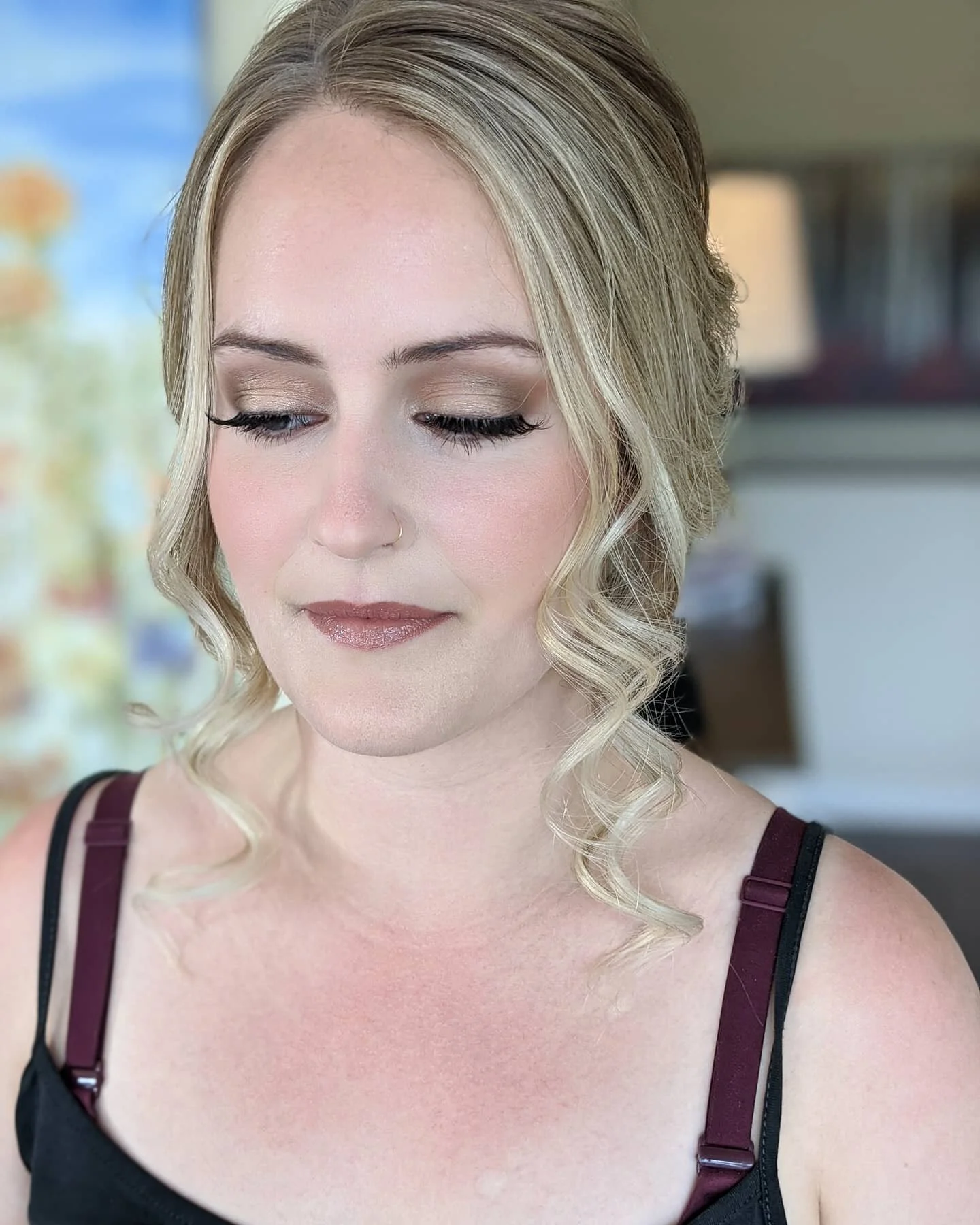 A woman with blonde hair styled in loose waves, wearing makeup with natural tones, a nose ring, and a black and maroon tank top, standing indoors with blurred background.